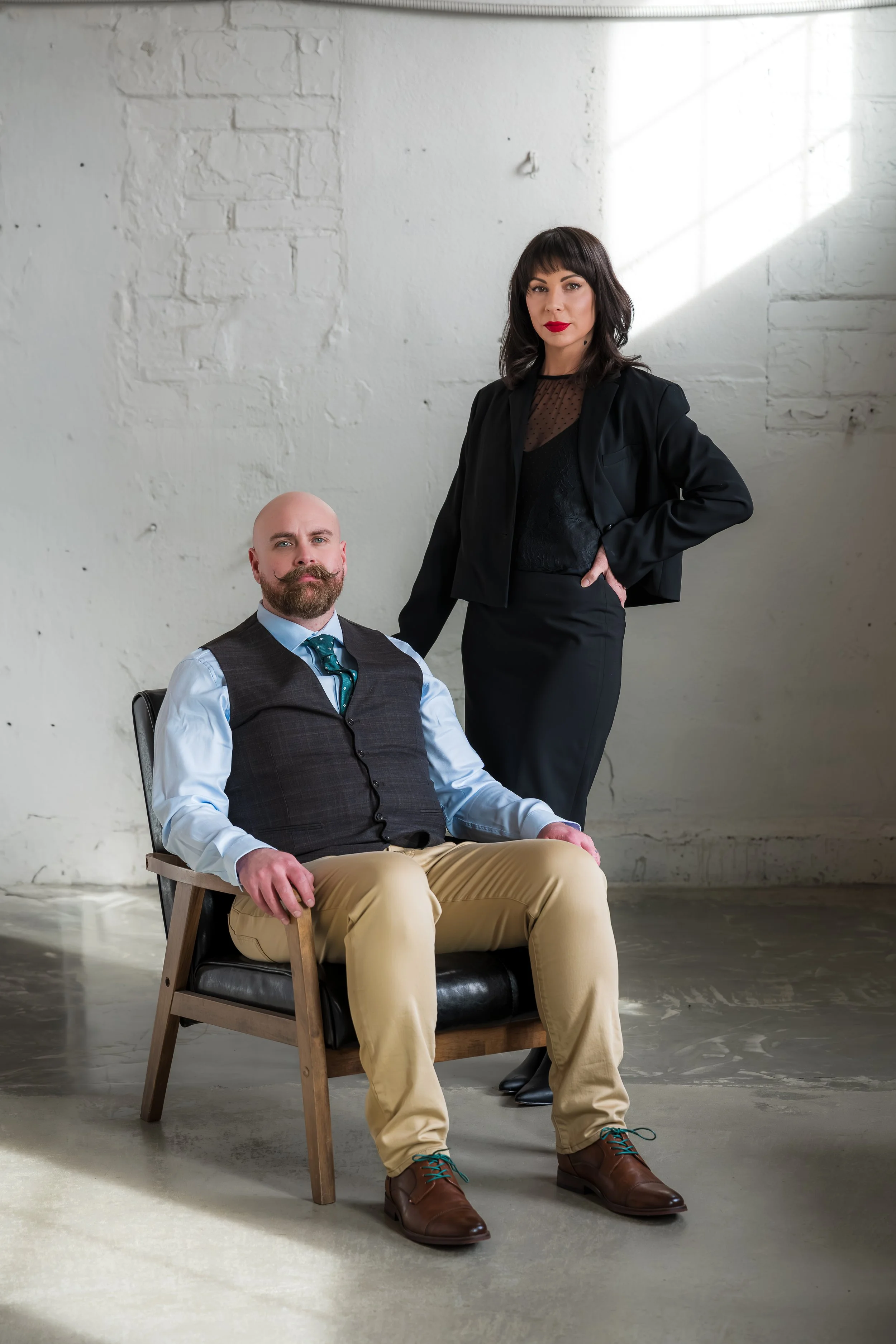 A man with a bald head, beard, and dressed in a light blue shirt, dark vest, and tan pants is seated in a wooden chair with black cushions, while a woman with dark hair, red lipstick, and dressed in a black blazer and skirt stands beside him with her hand on his shoulder. They are in an industrial-style room with white painted brick walls and concrete floor.