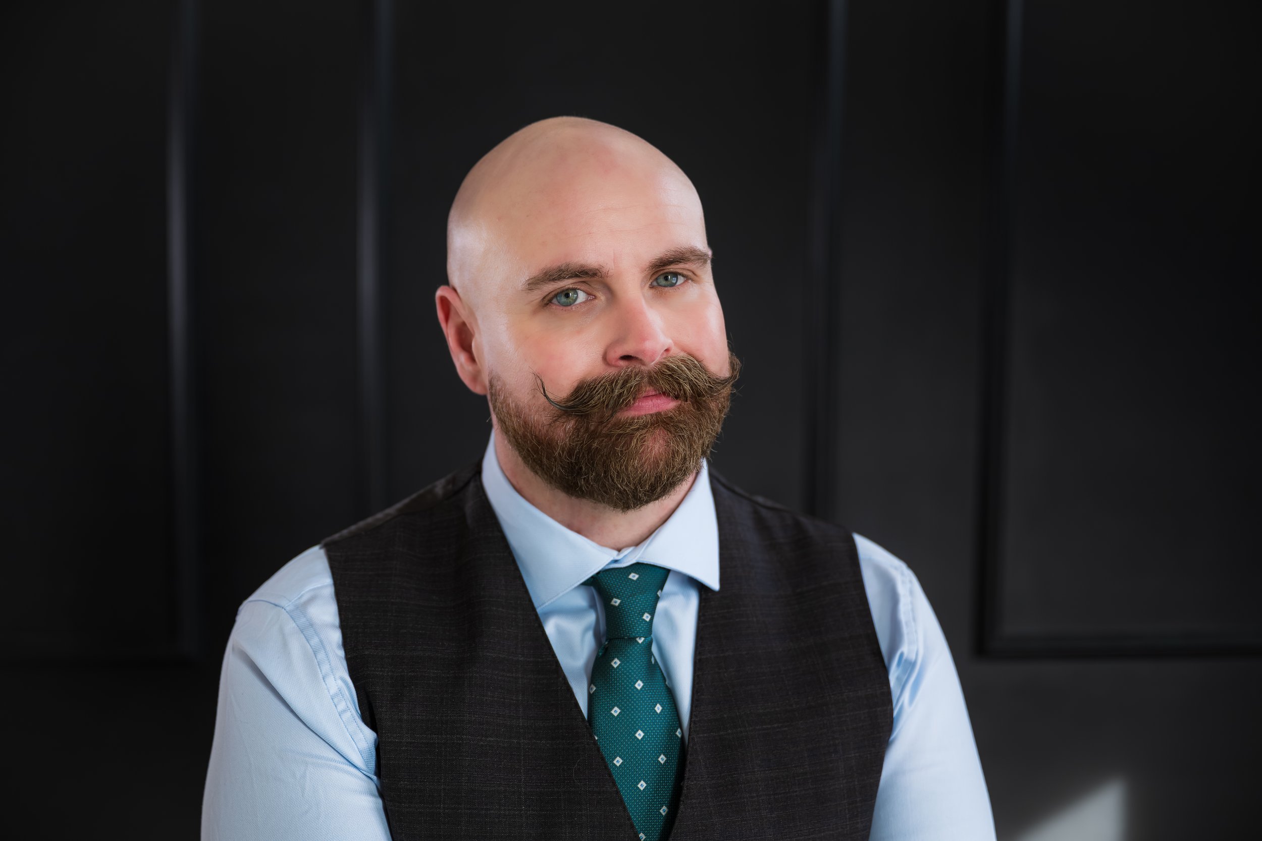Portrait of a bald man with a beard and mustache, wearing a dark vest over a light blue shirt and a teal tie with small white patterns, against a dark background.