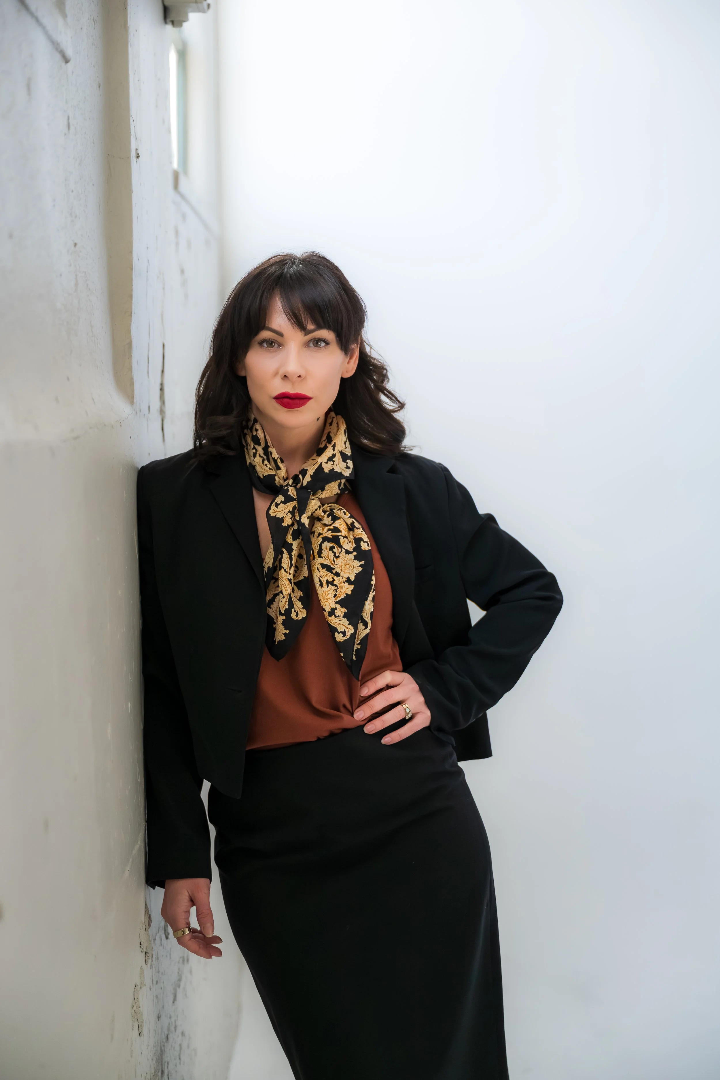 A woman dressed in business attire, with dark hair and red lipstick, leaning against a wall with a serious expression.