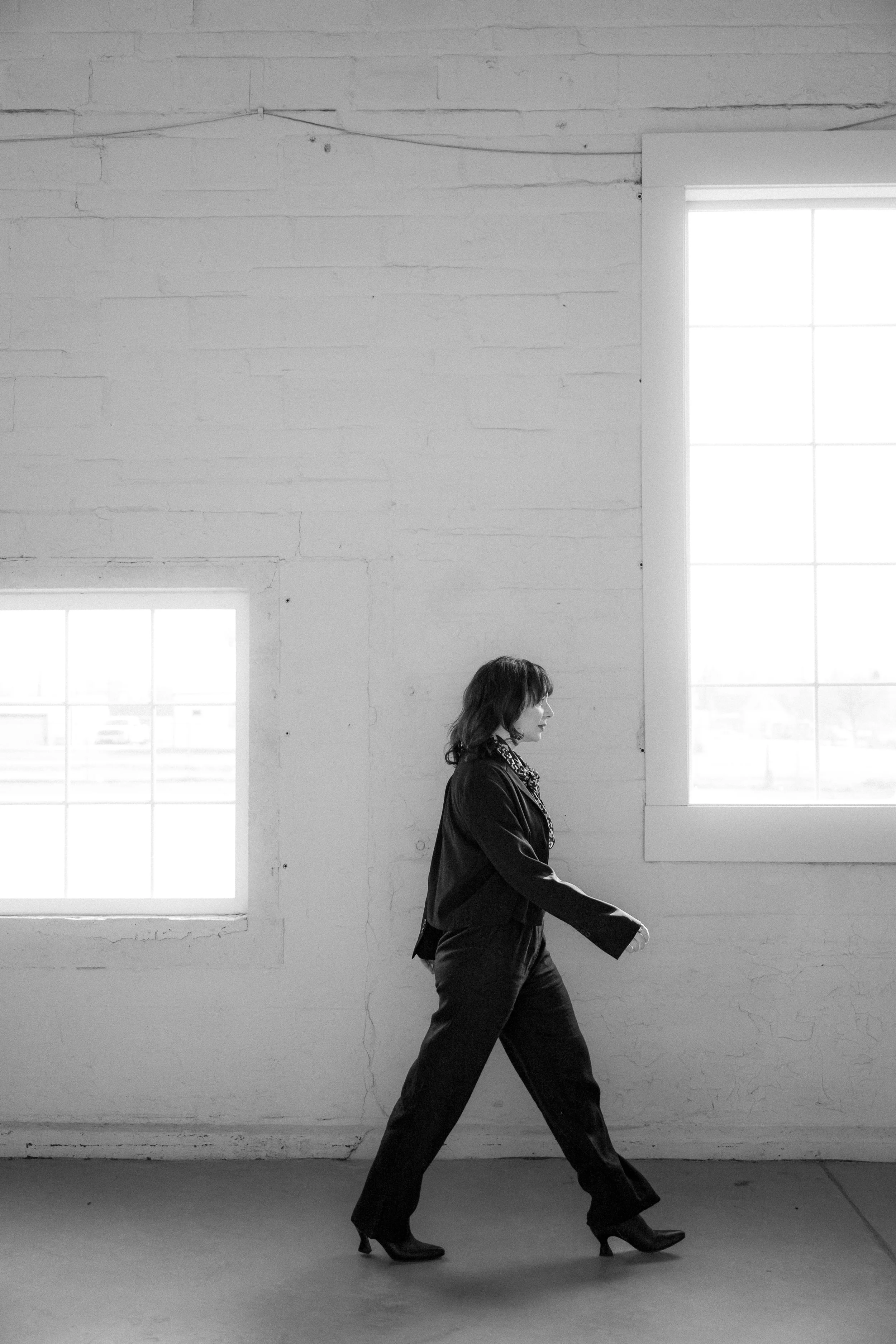 A woman walking indoors in front of large windows with natural light, in black and white.