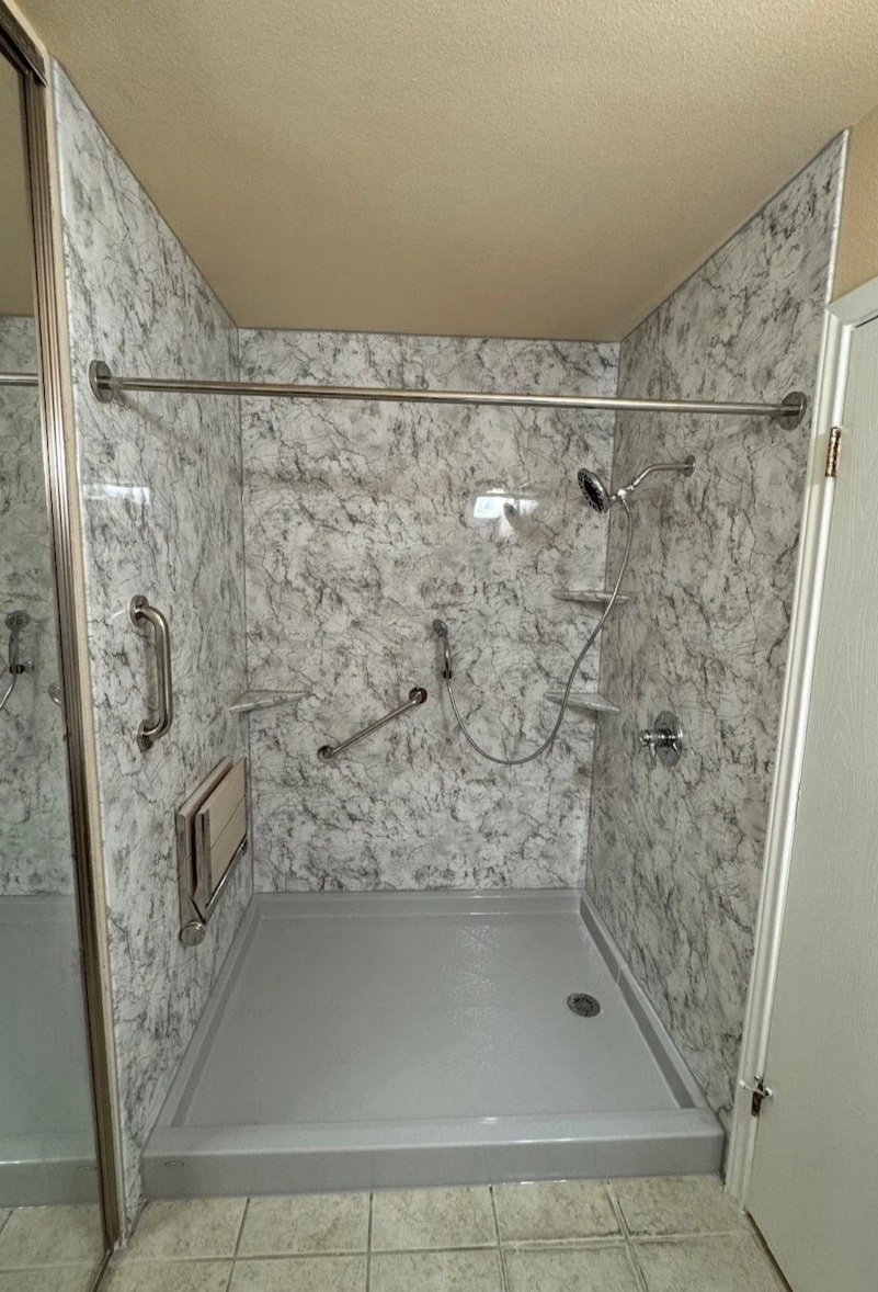 A walk-in shower with marble walls, a handheld showerhead, a shower curtain rod, safety grab bars, a fold down seat, and a drain in the center of the floor.