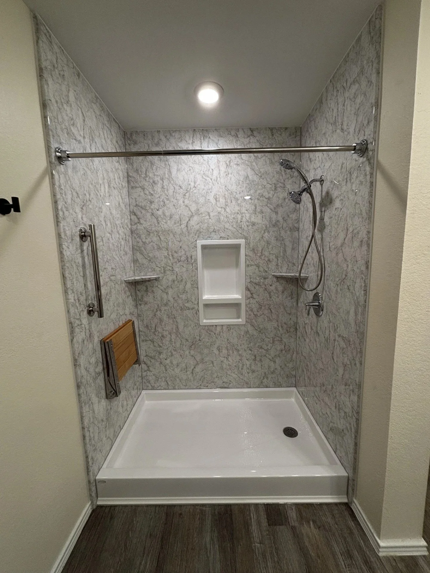 shower with marble walls, handheld showerhead, built-in shelves, grab bar, and wooden fold down seat