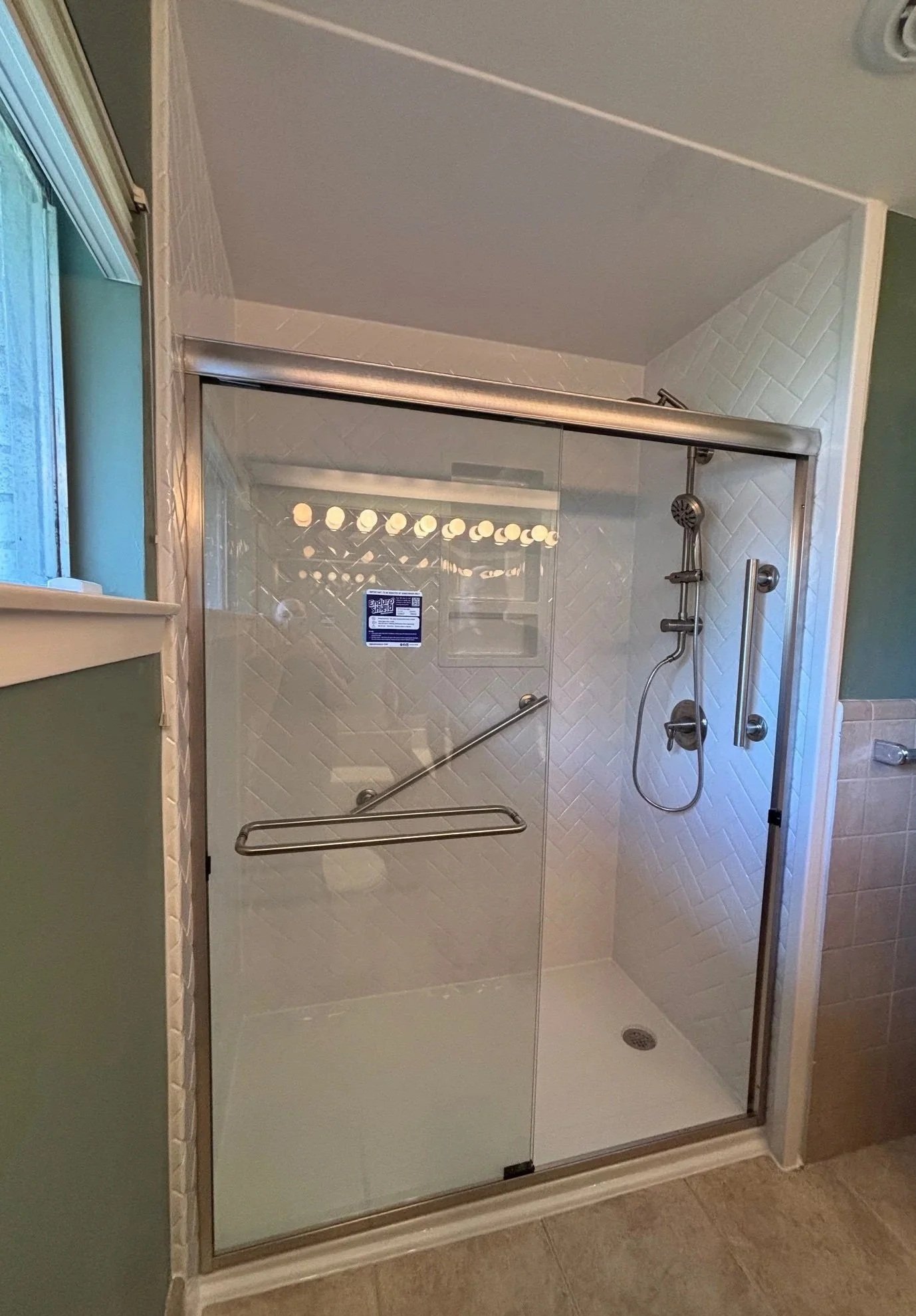 Glass shower enclosure with a sliding door, shower head, and grab bar inside a bathroom with taupe tiled floor.