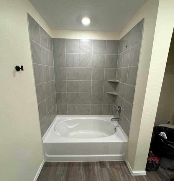 Bathroom with a bathtub, gray tiled wall, two corner shelves, and a ceiling light.