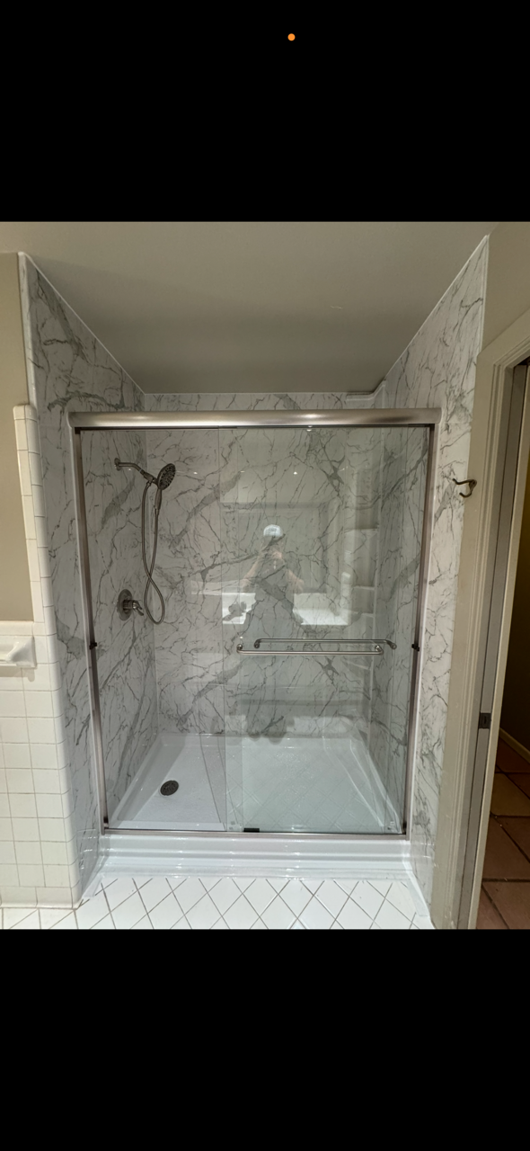 A walk-in shower with marble walls and a glass door, featuring a handheld showerhead and a towel bar.