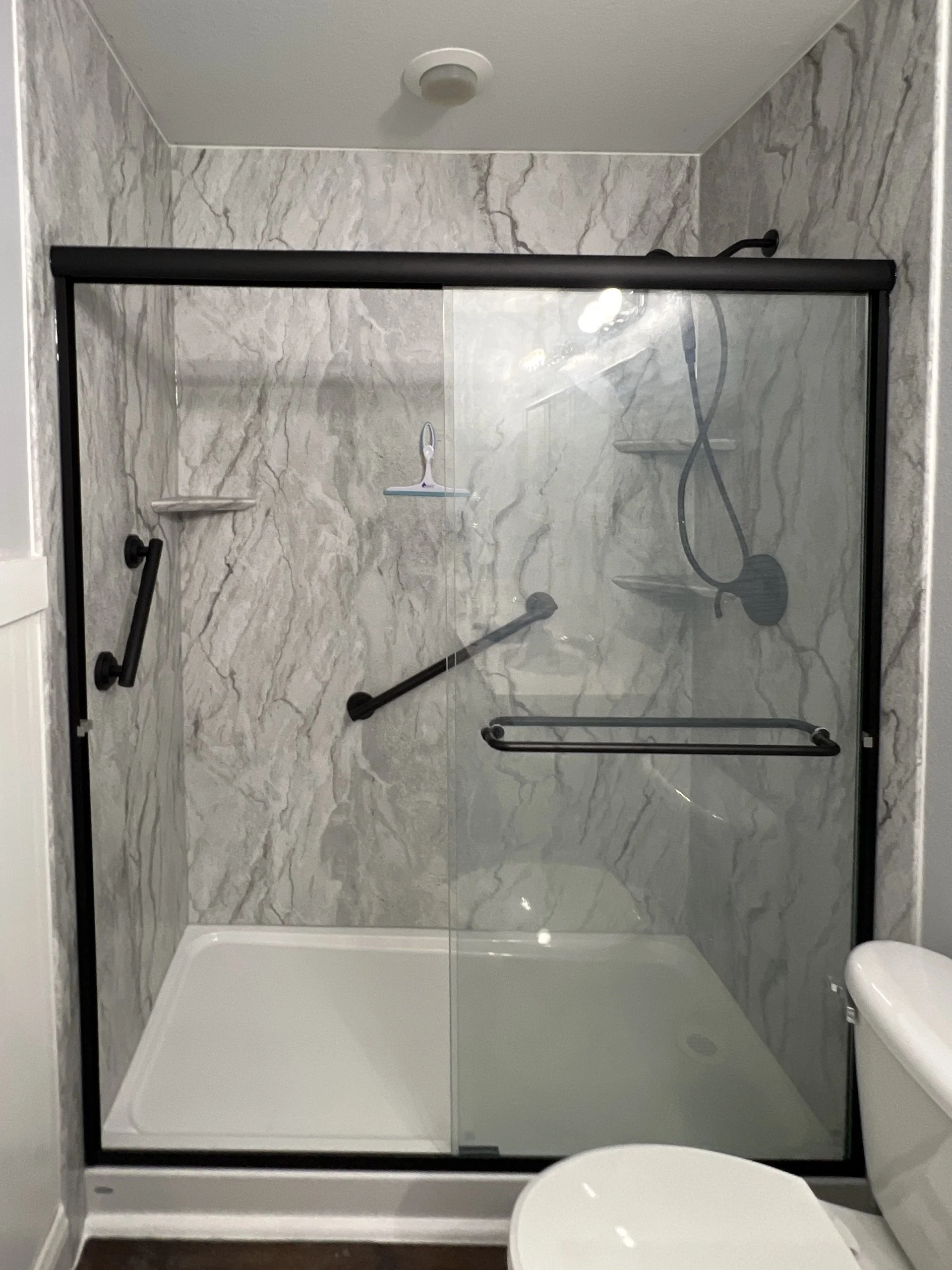 Acrylic shower, gray marble walls, glass doors, shower head, and small corner shelves.