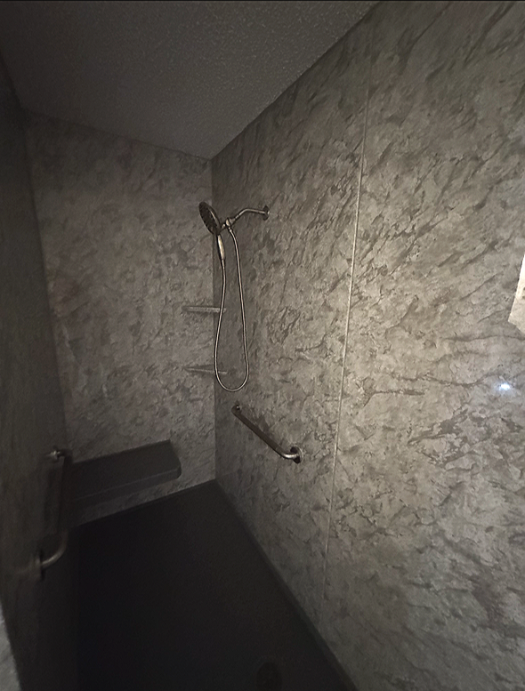 A shower area with gray marble walls, a handheld showerhead, a grab bar, and a fold-down seat.