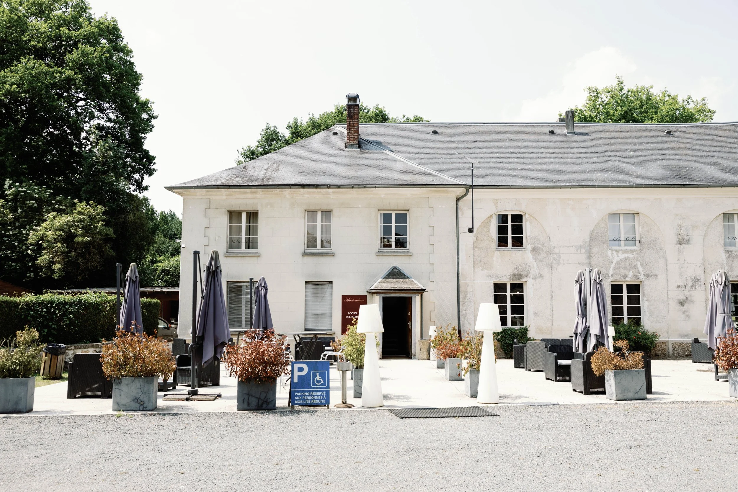 Exterior of a historic white building with outdoor seating area, umbrellas, potted plants, and a parking reserved sign, surrounded by trees.