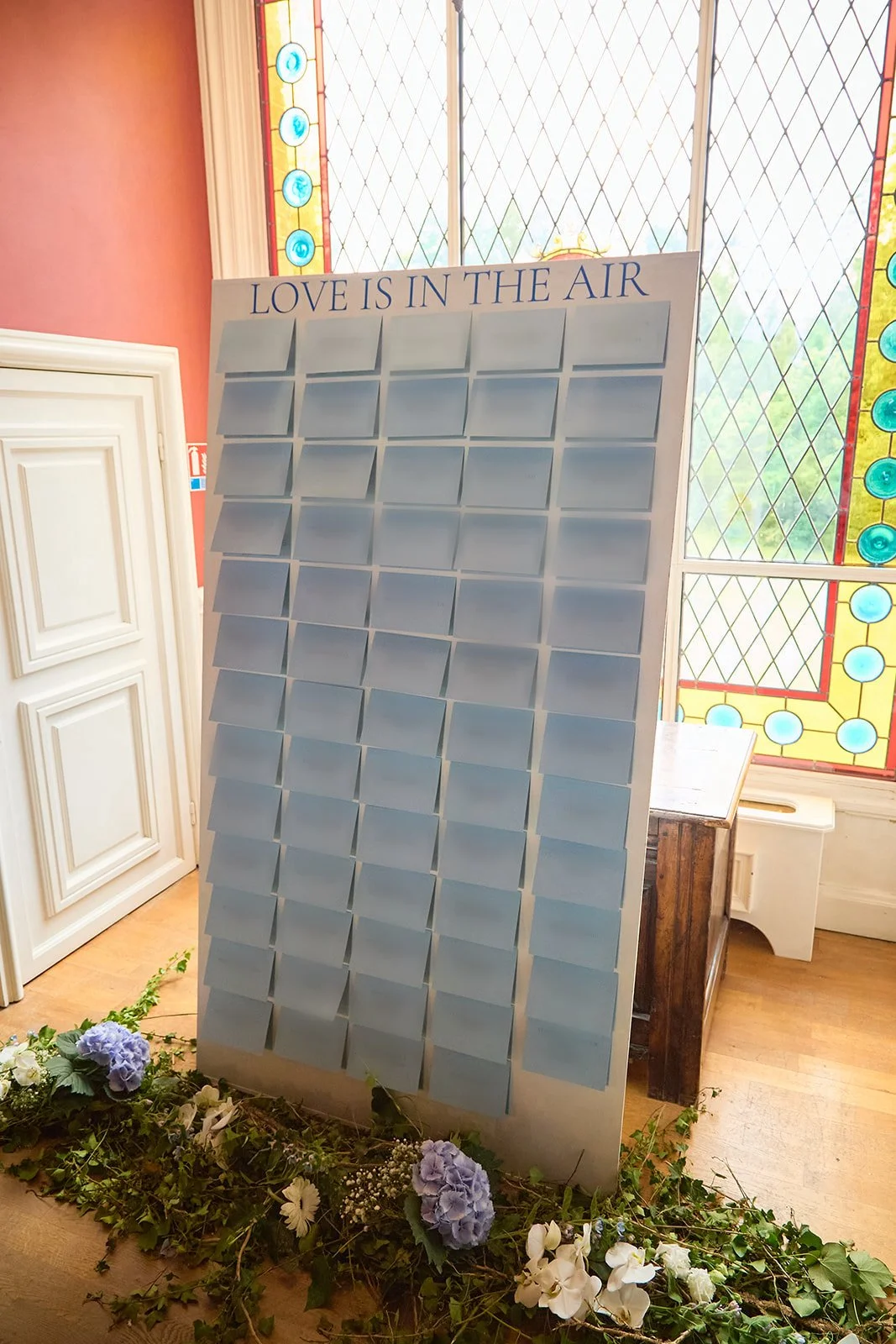 Large blank board with the words "LOVE IS IN THE AIR" at the top, surrounded by flowers and greenery at the base, set in a room with stained glass windows.