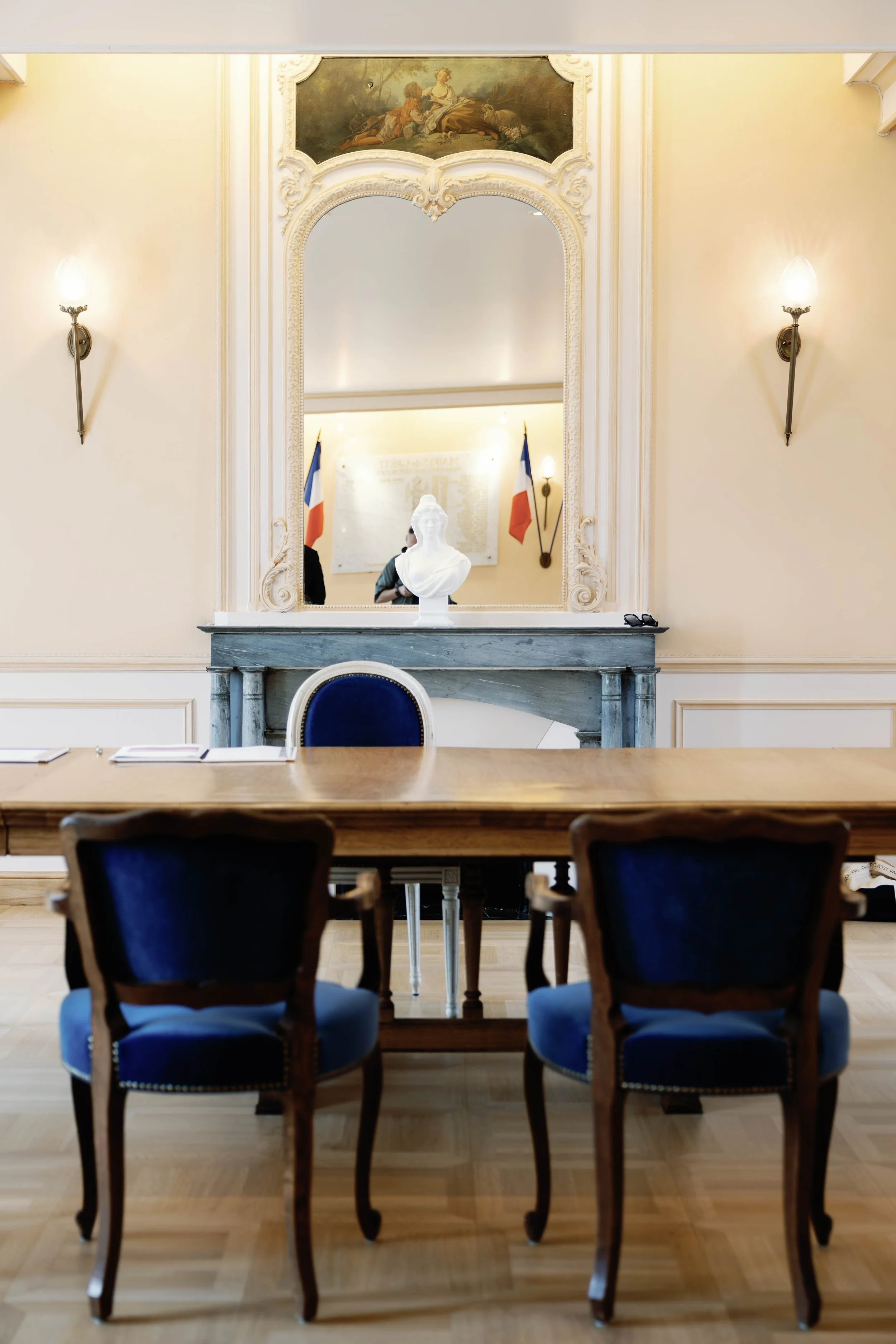 A formal meeting room with a large wooden table, three chairs with blue velvet cushions, a mirror above a gray marble fireplace, a bust sculpture, two French flags, and a painting of a historical scene on the wall.