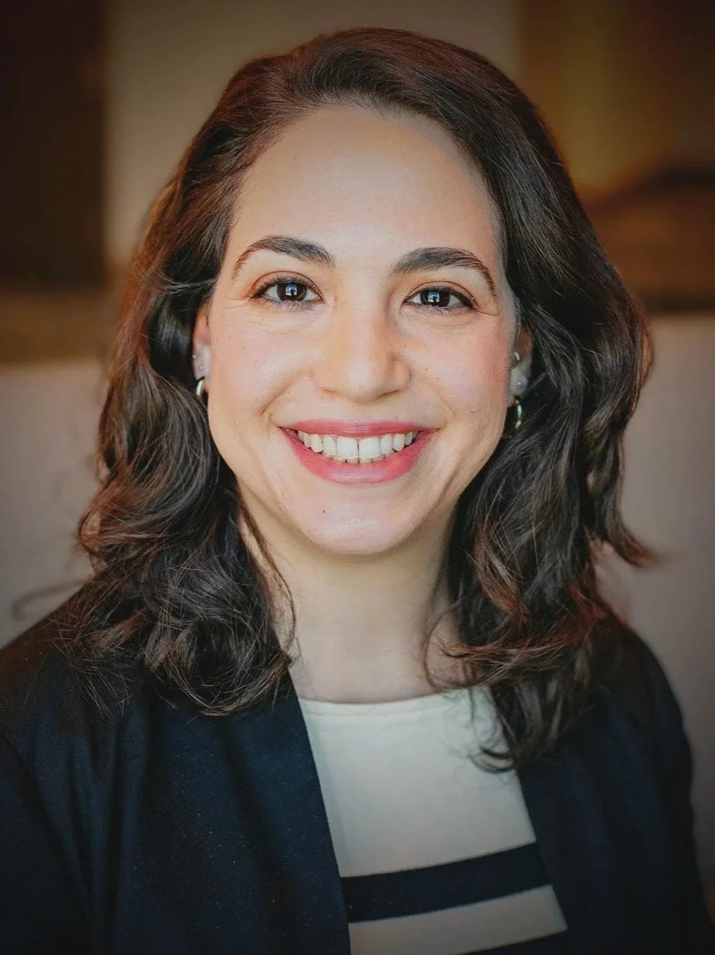 A woman with wavy dark hair smiling at the camera, wearing earrings and a black blazer over a white top with black stripes.