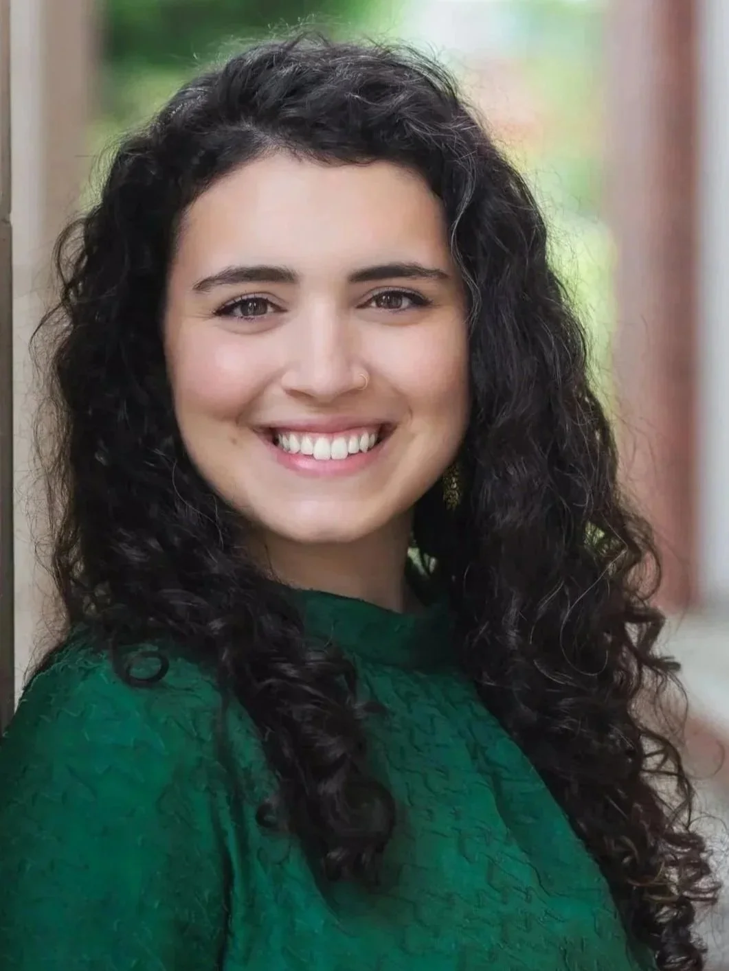 Portrait of a woman with long, curly dark hair, smiling, wearing a green top, outside with blurred background.