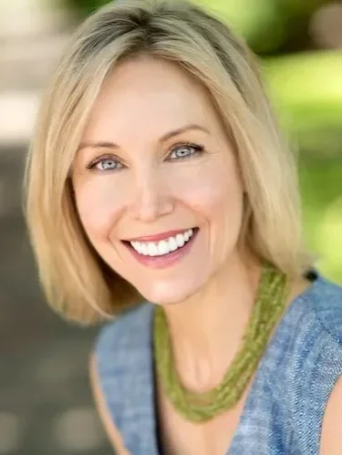 Smiling woman with blonde hair, wearing a blue top and green necklace, outdoors with blurred green background.
