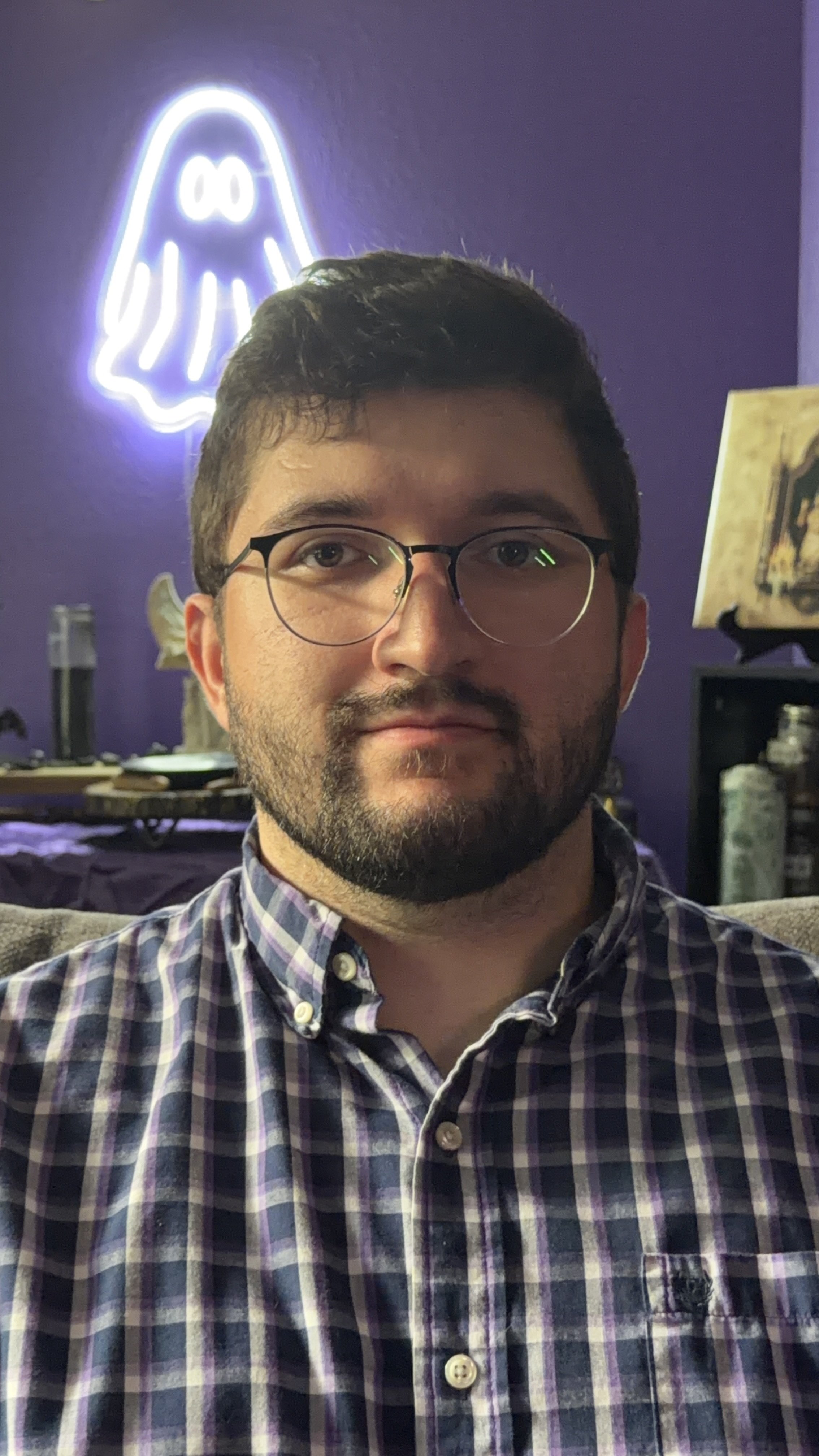 A young man with glasses and a beard taking a selfie in a room with purple walls, featuring a neon ghost sign and various decorative items in the background.