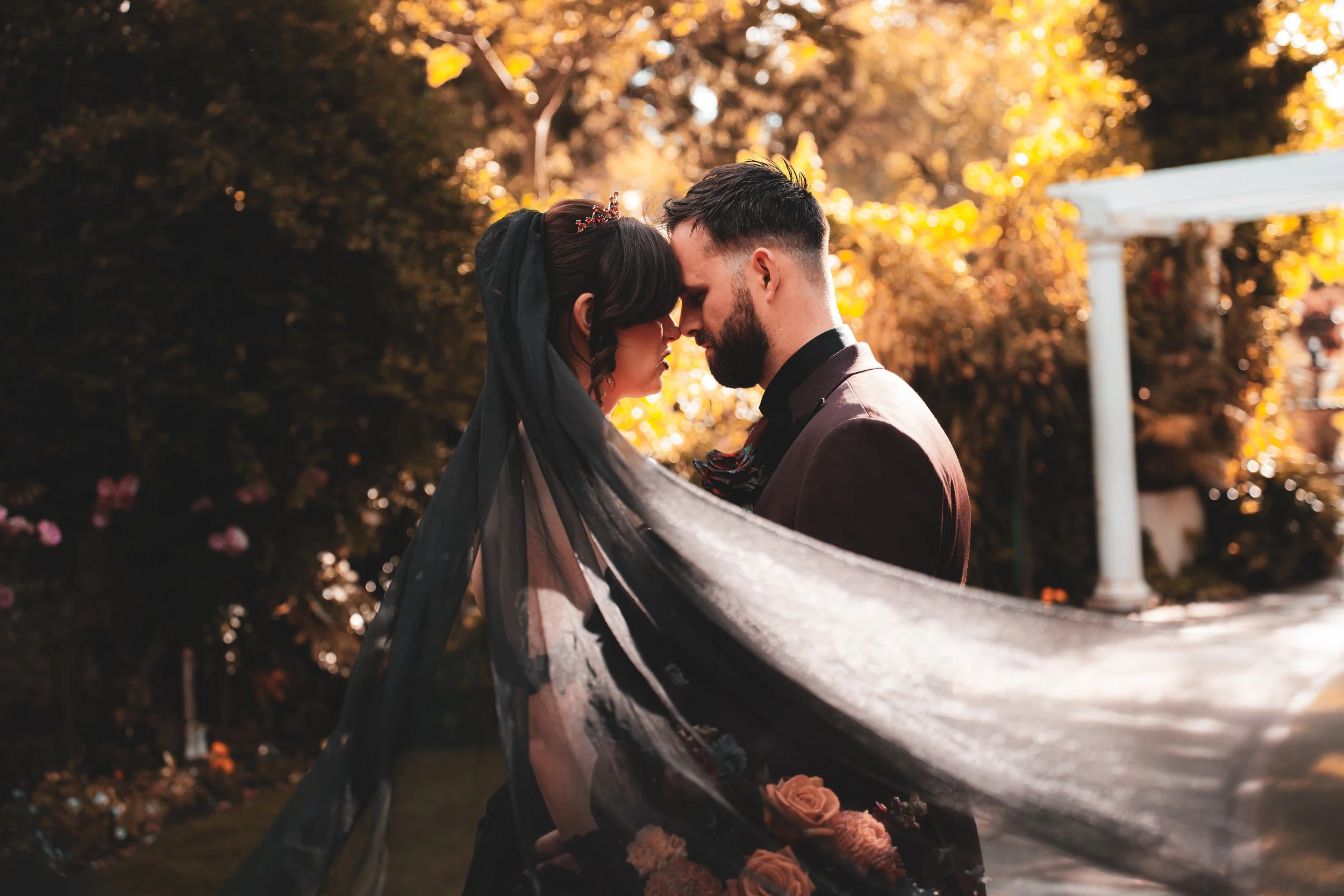 A bride and groom face each other with foreheads touching, outdoors with autumn-colored foliage in the background. The bride wears a black veil and floral gown, and the groom in a dark suit.
