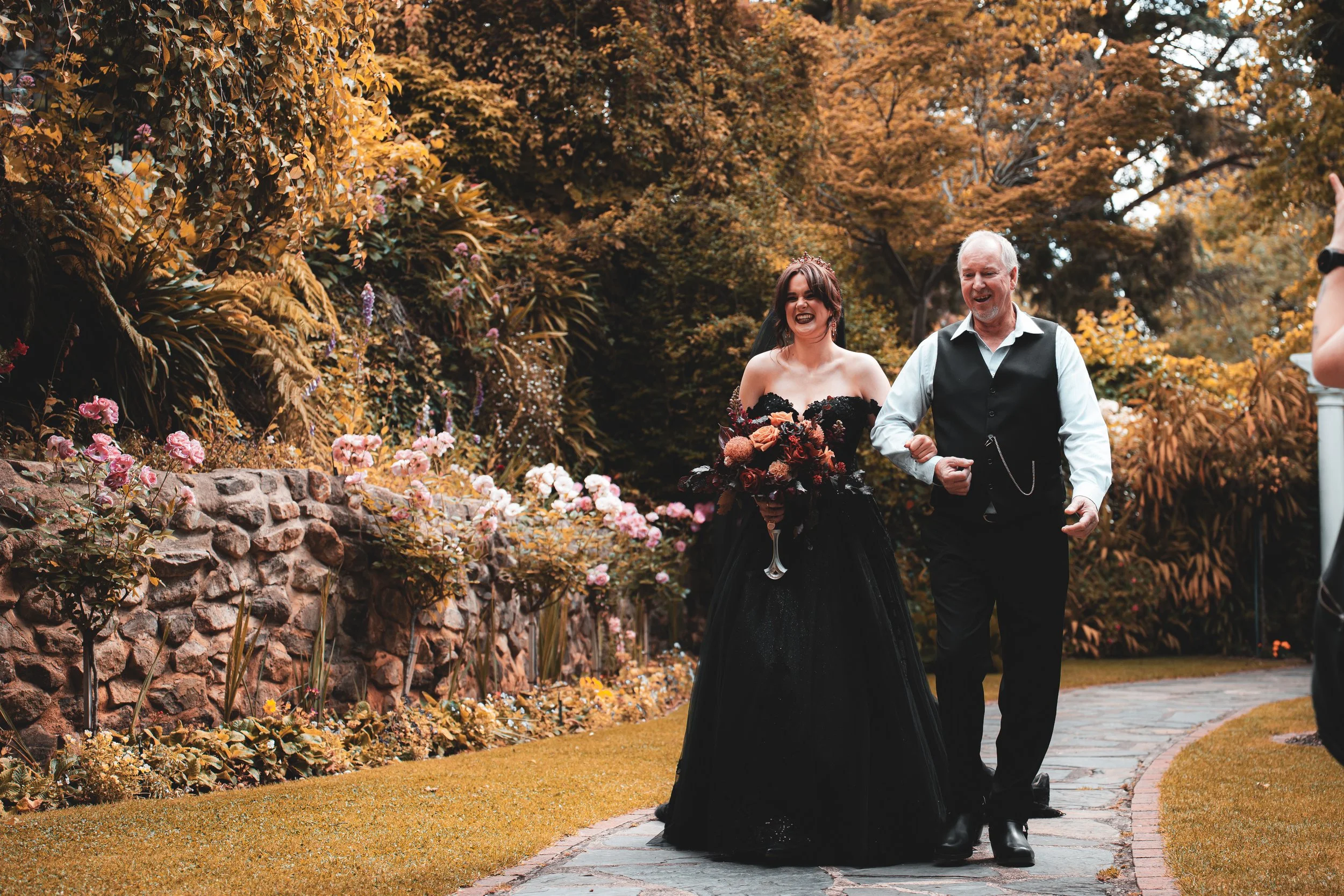 A woman in a black gown holding a bouquet of flowers walking arm-in-arm with a man in a vest and white shirt, smiling, in a garden with fall foliage.