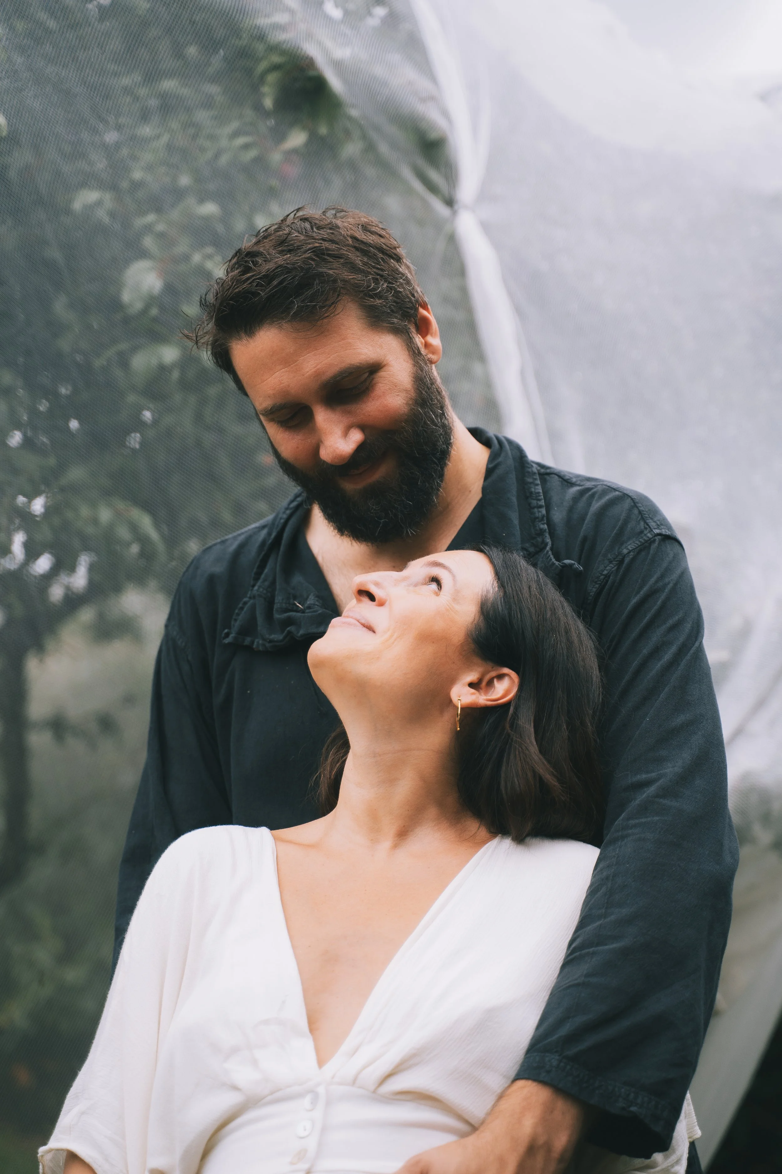 A man and woman sharing a tender moment outdoors, with the man embracing the woman from behind and both looking at each other with affection.
