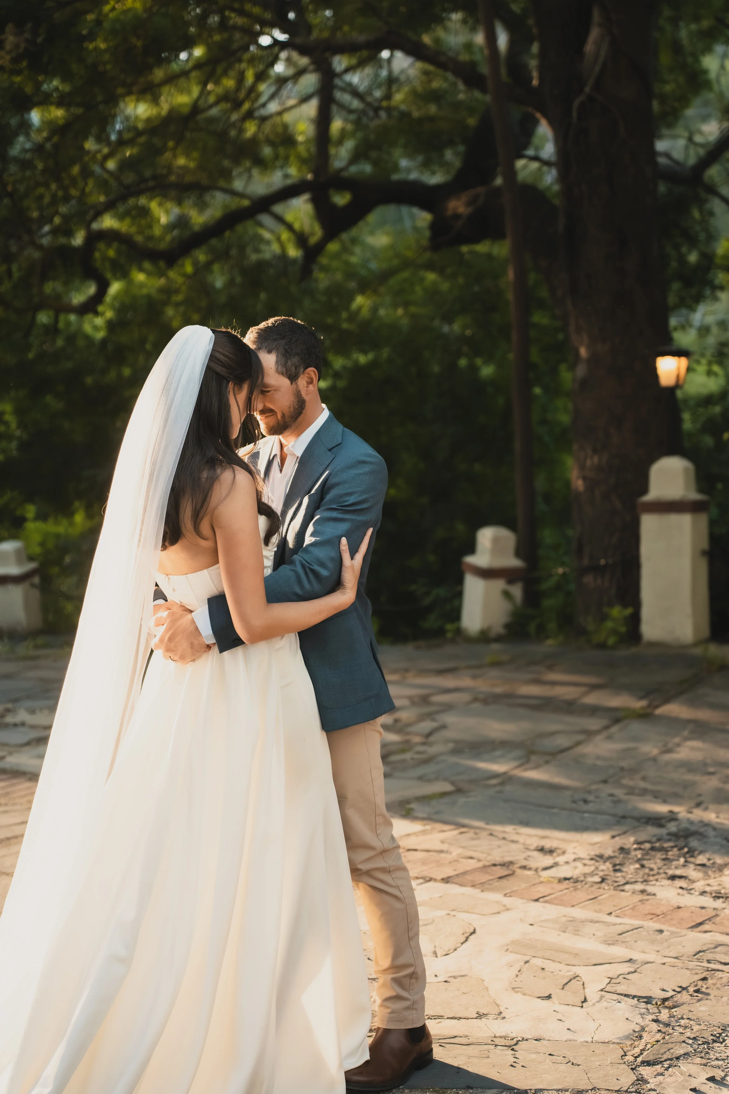 A bride and groom share a close, intimate moment outdoors during their wedding, standing on a stone pathway with lush green trees in the background.