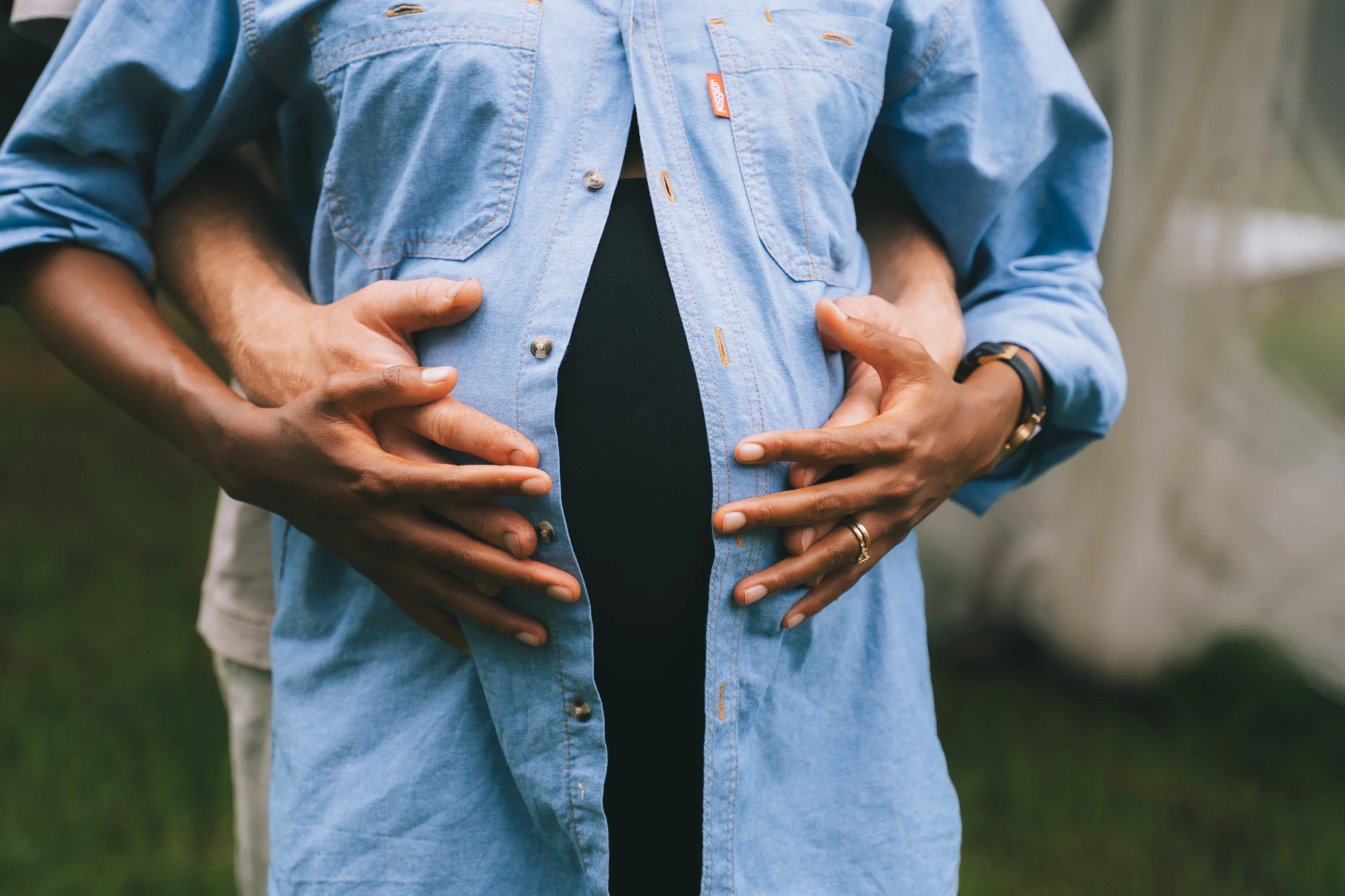 Four hands, belonging to two people, are placed on a pregnant woman's abdomen, with the woman wearing a light blue denim shirt unbuttoned over a black top, against a blurred outdoor background.