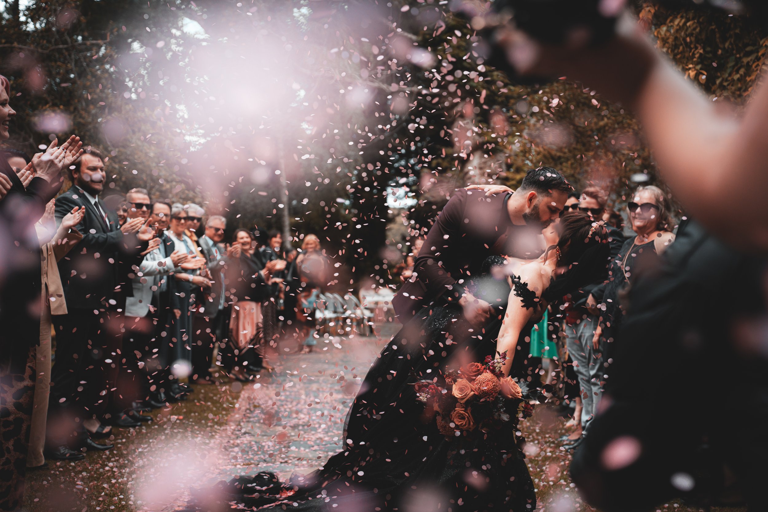 A newlywed couple kisses while walking through a shower of pink and white confetti, surrounded by family and friends celebrating at an outdoor wedding ceremony.