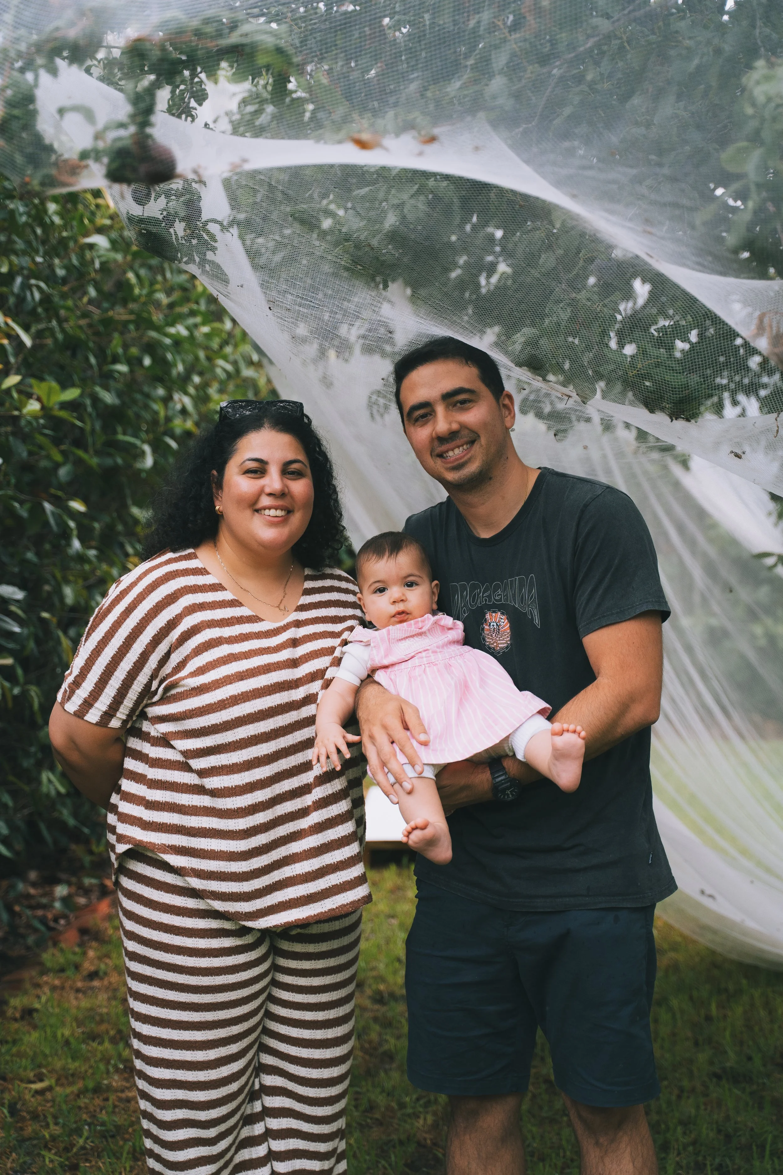 A family of three smiling outdoors, holding a baby girl dressed in pink, under a plastic sheet canopy among greenery.