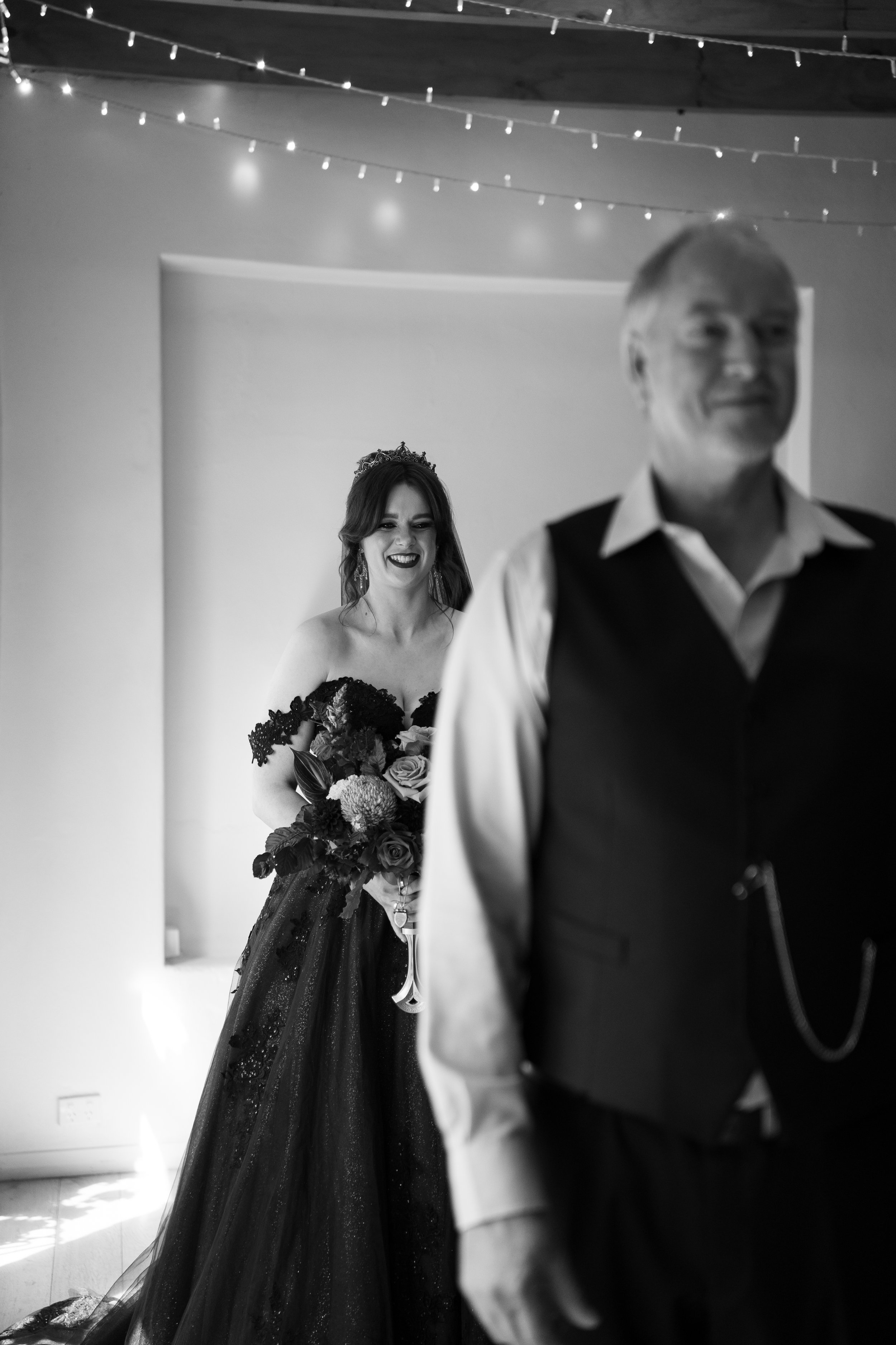 A bride smiling while holding a bouquet of flowers, standing behind a man who is slightly blurred in the foreground, with string lights overhead in a wedding reception.