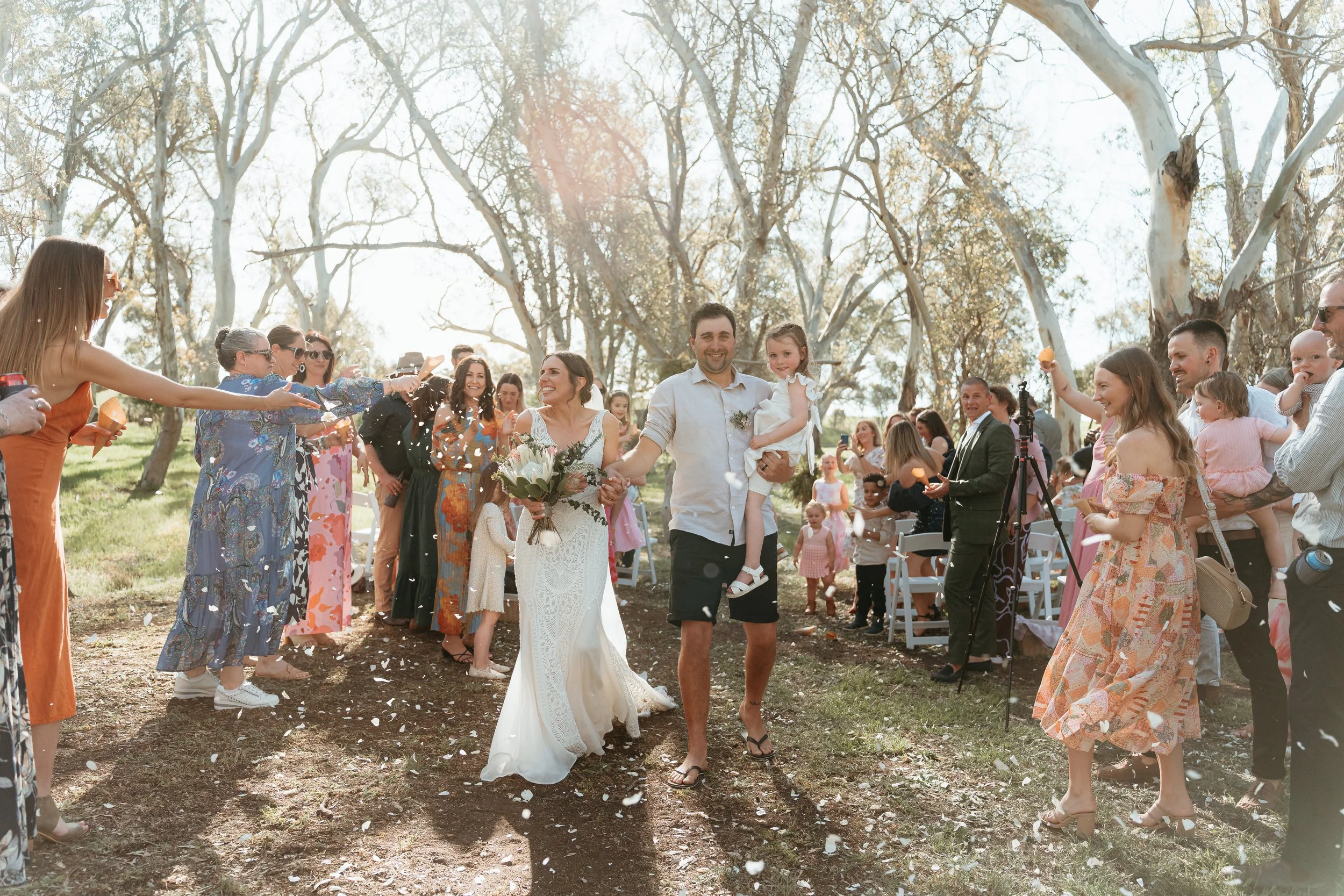 A wedding celebration outdoors with guests throwing flower petals as a bride and groom walk down the aisle, surrounded by family and friends in sunlight and trees.