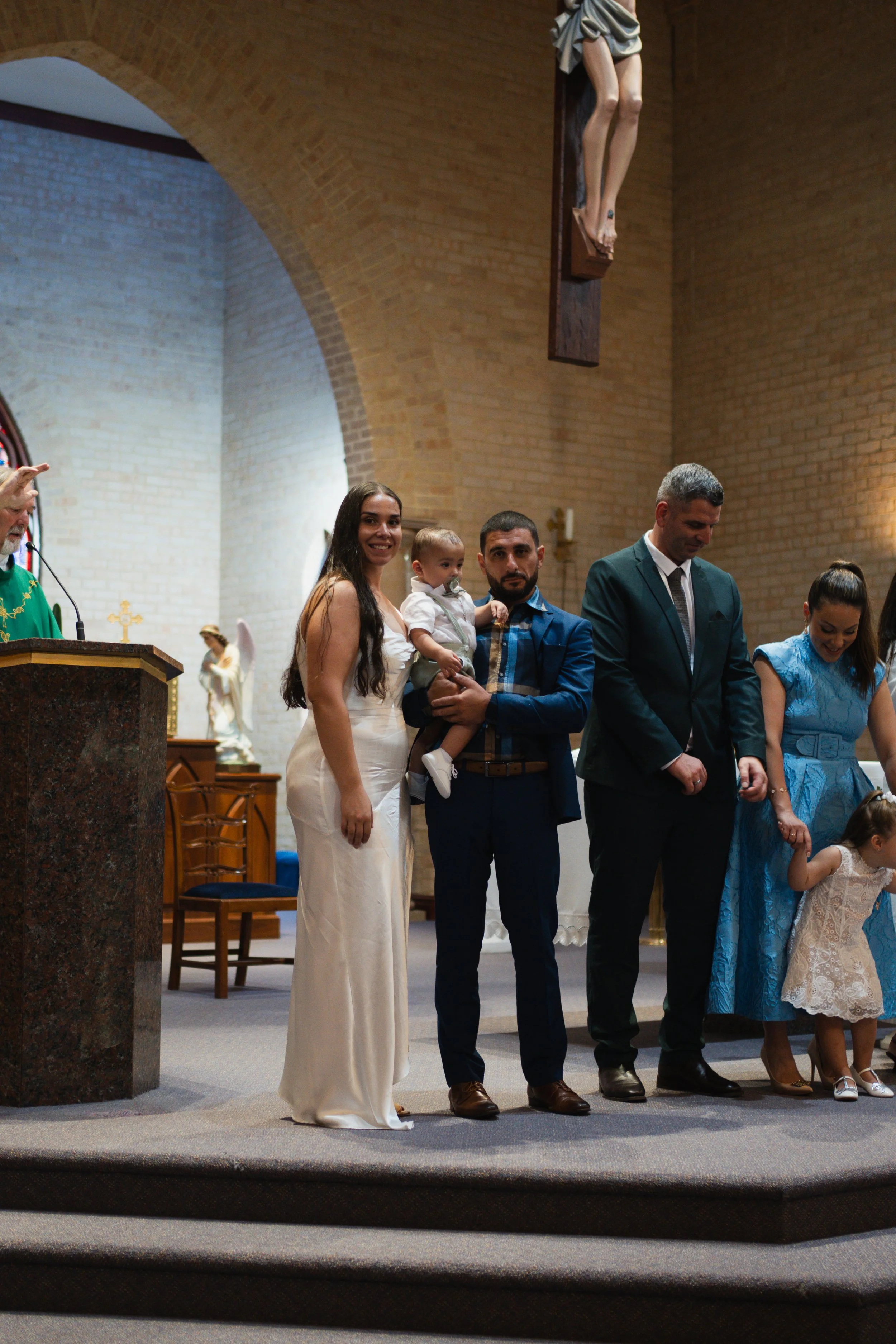 A group of people dressed in formal attire standing inside a church. A woman in a white dress, a man holding a child, a man in a suit, and a woman with a young girl, with a priest at the altar.