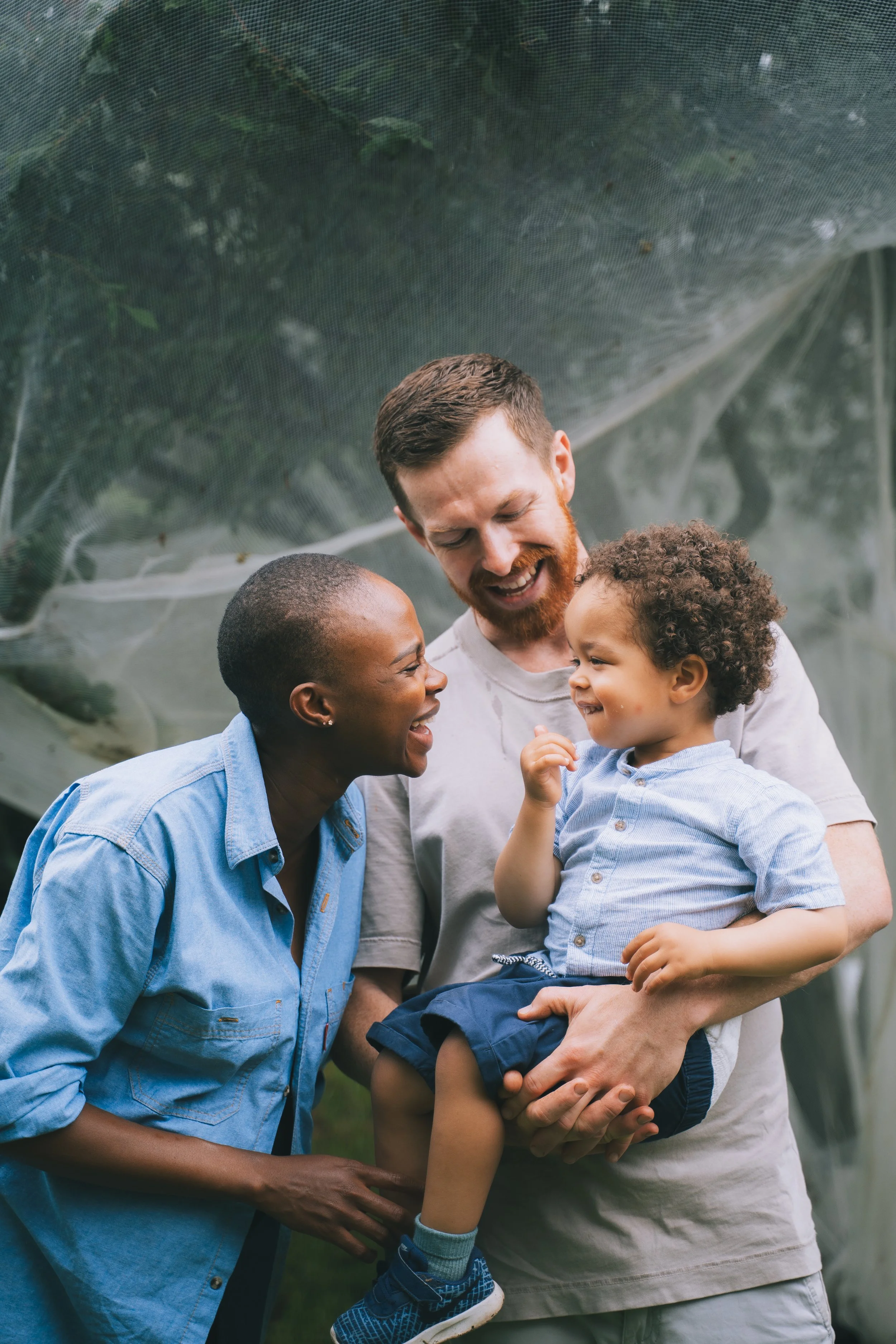 A happy family of three outdoors, with a woman and man smiling at a young boy in the man's arms. The boy is smiling and looking at the woman, all smiling and enjoying a joyful moment.
