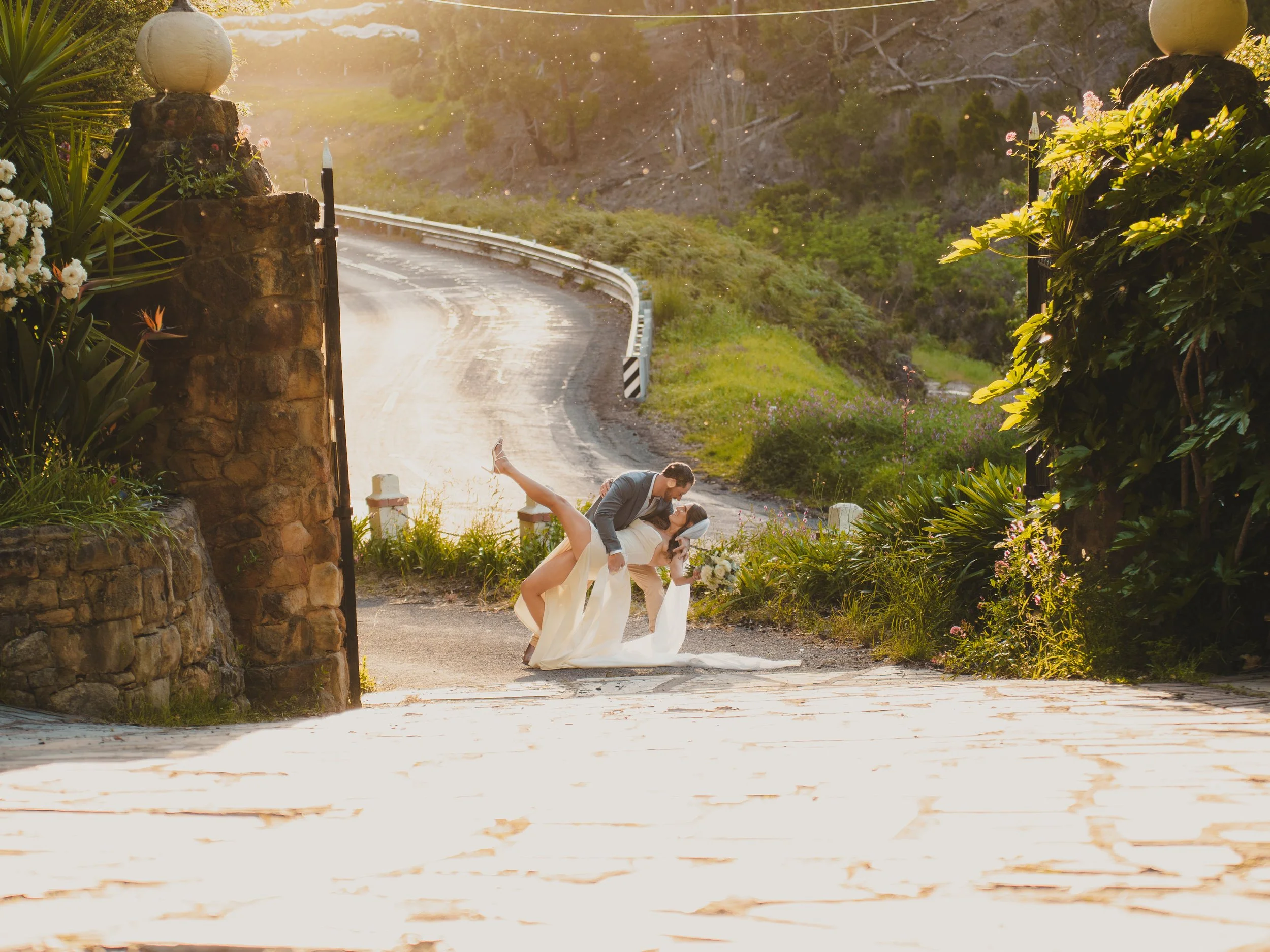 A newlywed couple shares a romantic kiss in a scenic outdoor setting during sunset, with the groom dipping the bride while holding her waist and the bride holding a bouquet of flowers.