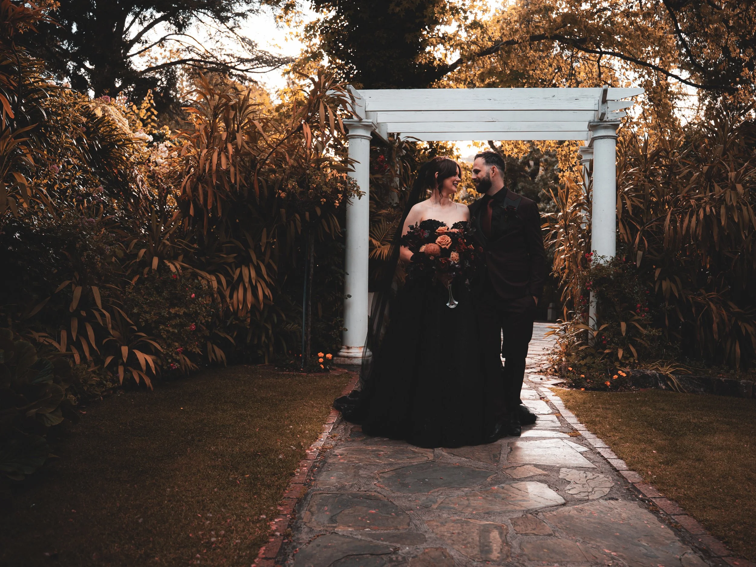 A couple in formal attire standing close together under a white garden archway, holding a bouquet of flowers, surrounded by lush garden foliage during sunset.