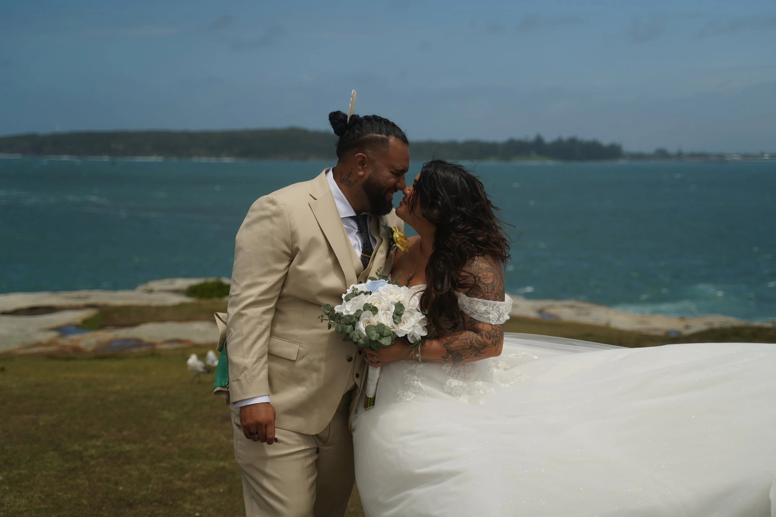 A newlywed couple sharing a kiss by the water, with the bride holding a bouquet of white roses and the groom dressed in a light tan suit, near a rocky shoreline with the ocean in the background.