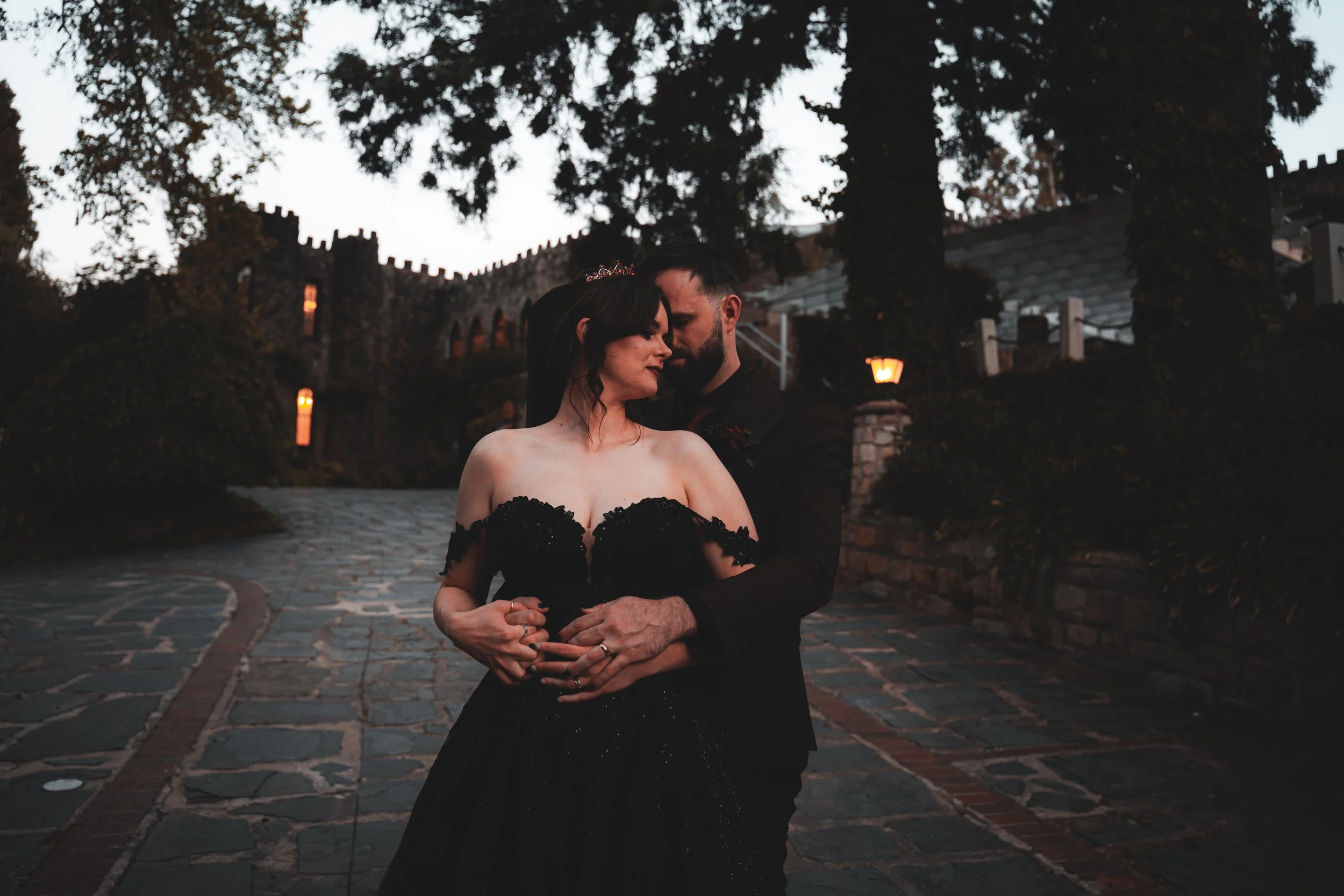 A romantic couple dressed in formal wear, embracing on a cobblestone pathway in an outdoor setting during dusk, with trees and castle-like structures in the background.