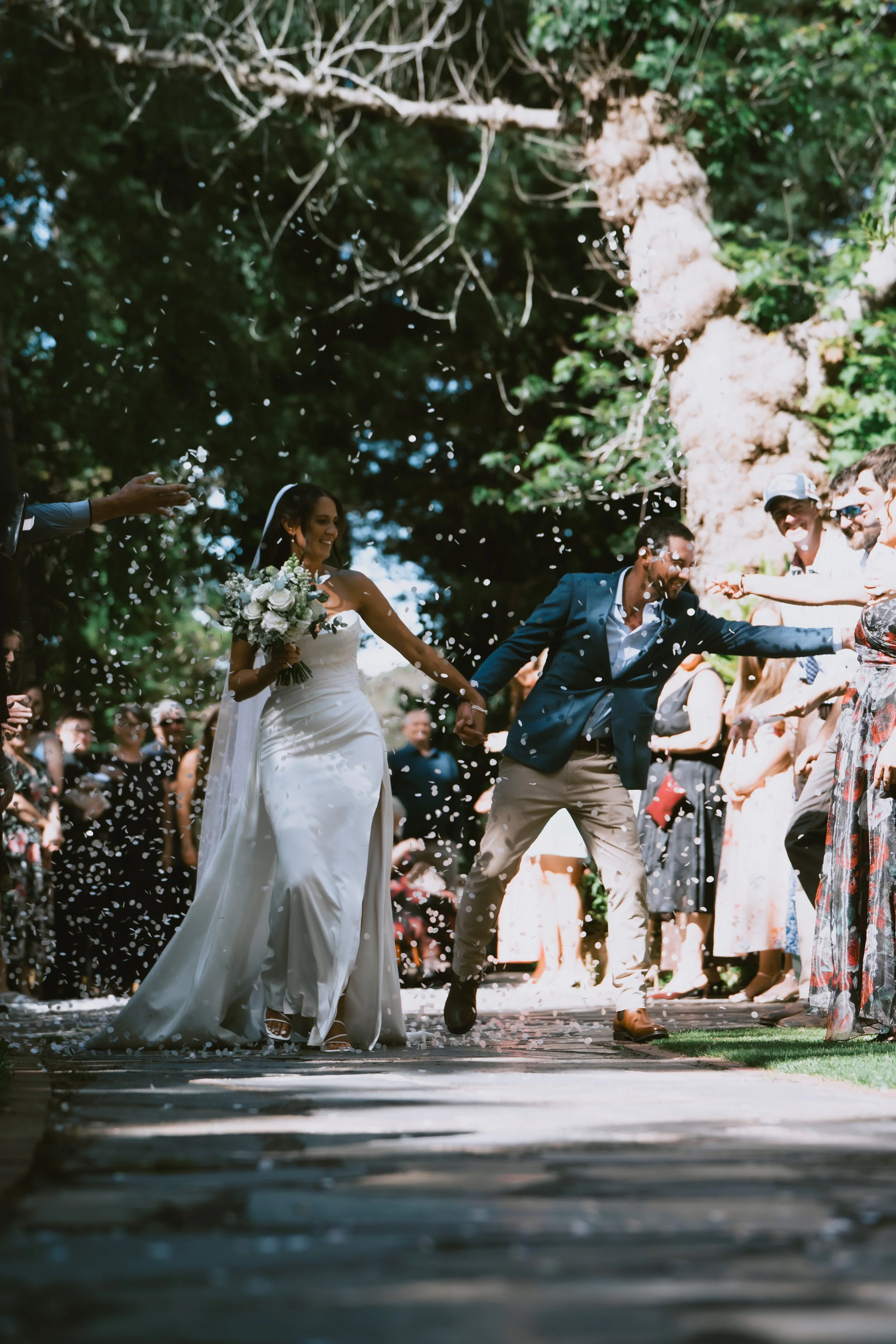 Bride and groom happily walking hand in hand through a shower of rice or confetti during their outdoor wedding ceremony, surrounded by friends and family.