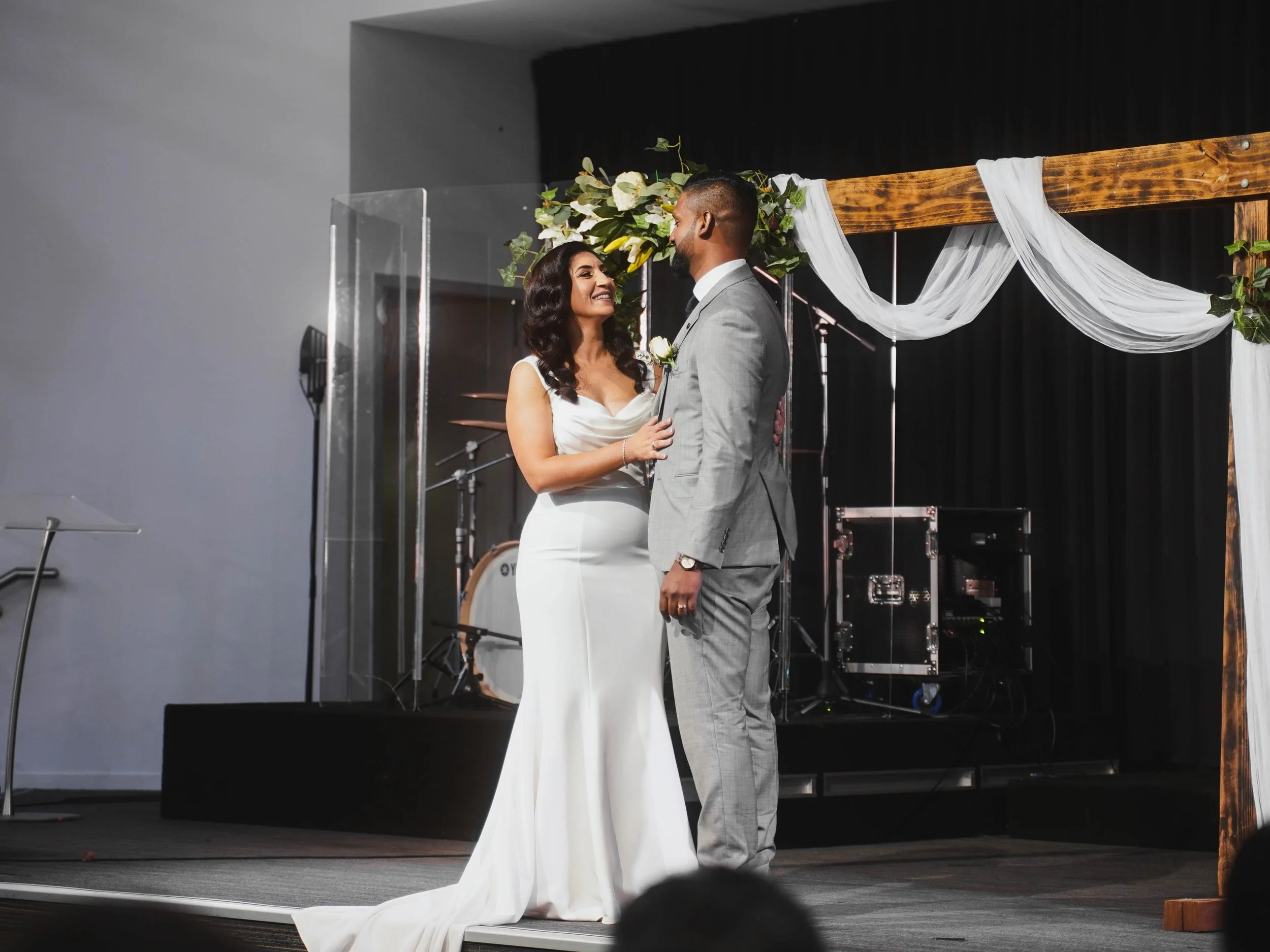 A bride and groom exchange vows during their wedding ceremony on a decorated stage with an arch, flowers, and drapery.