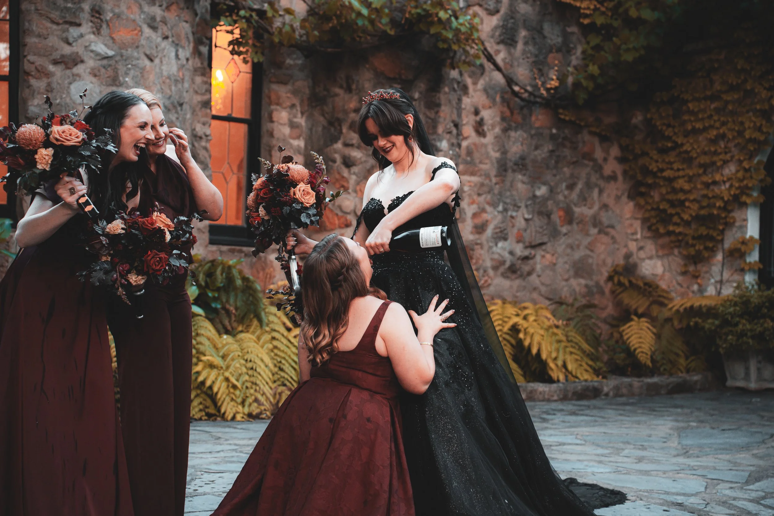 A bride in a black wedding dress with lace and tulle is being poured champagne on by a woman kneeling in a magenta dress. Two women in dark red bridesmaid dresses with bouquets are smiling and laughing, standing in front of a stone wall with windows 