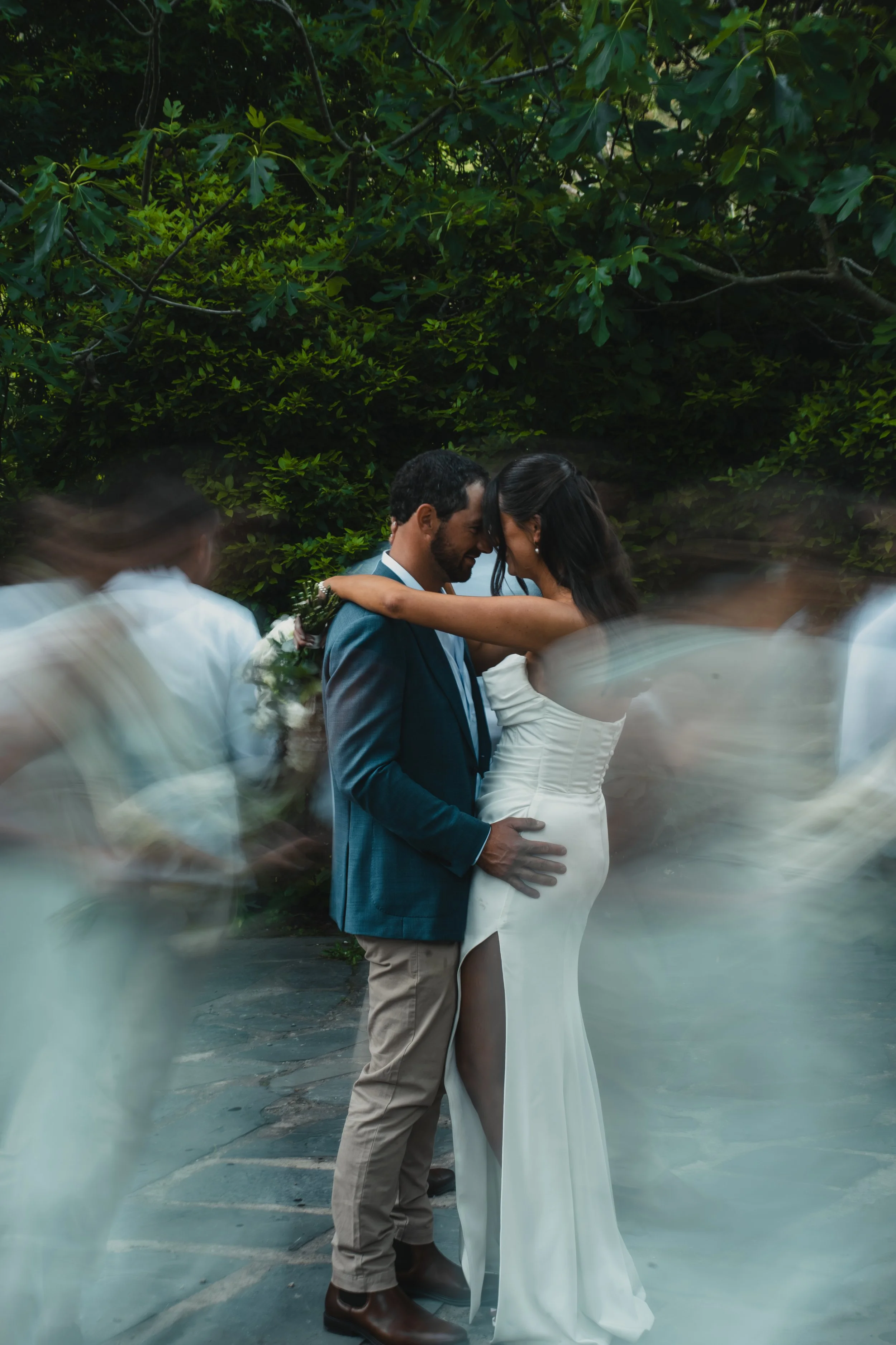 A couple having a romantic moment, standing close with foreheads touching, surrounded by blurred wedding guests at an outdoor wedding.