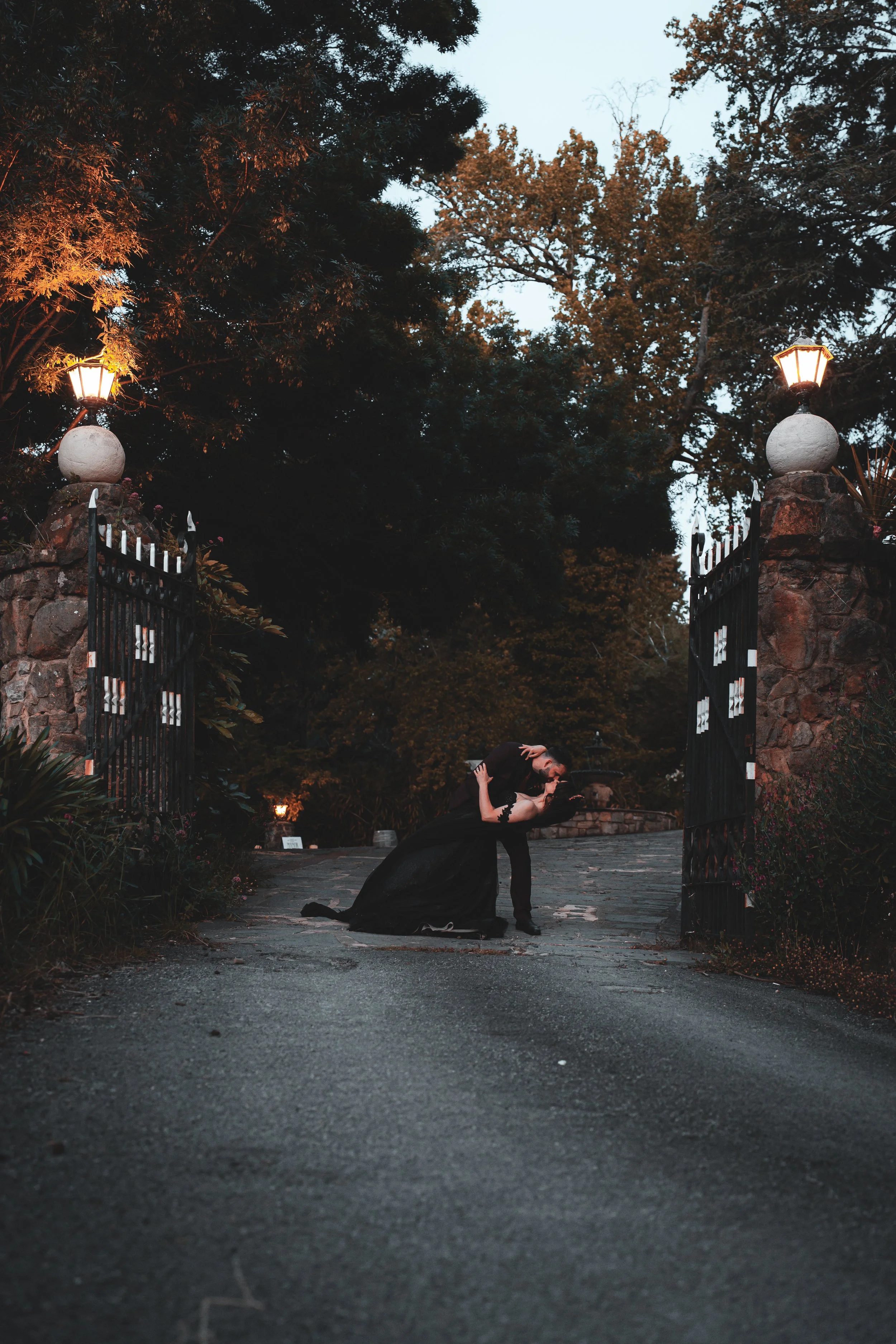 Person dressed in black kneeling on the driveway with a dramatic pose at an open gate decorated with stone and ironwork, illuminated by street lamps, surrounded by trees at dusk.