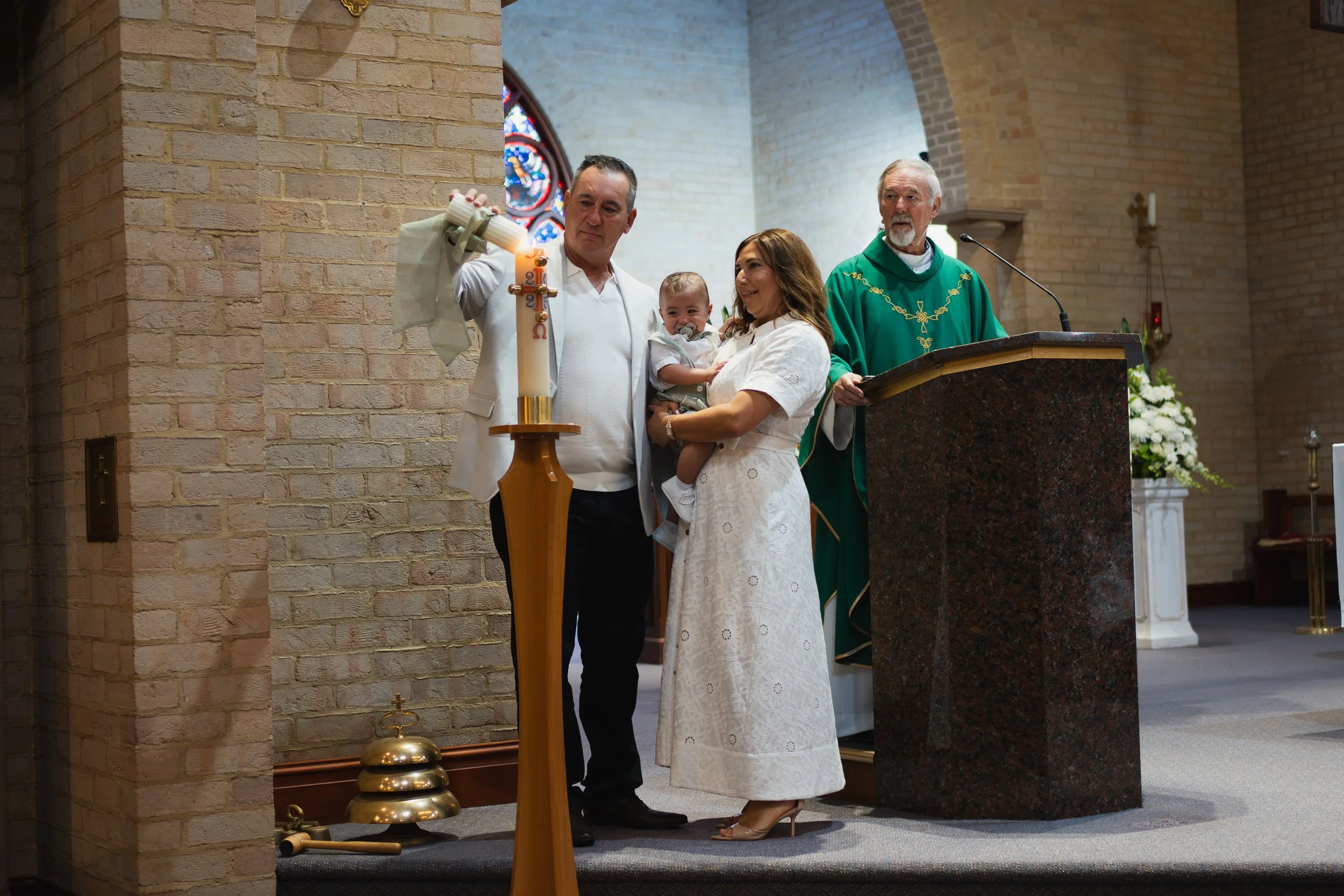 A family standing at an altar during a baptism ceremony inside a church, with a priest in green robes officiating.