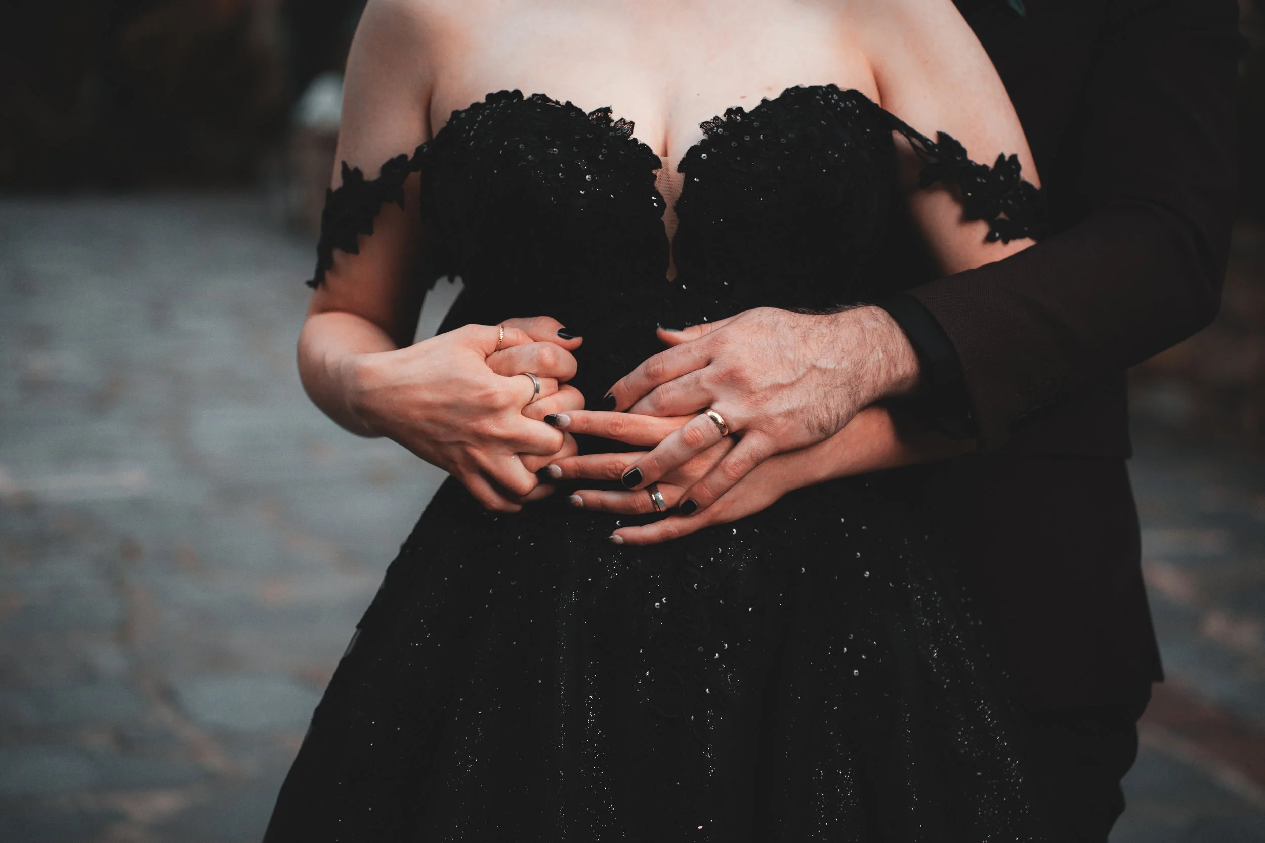 Close-up of a couple holding hands with wedding rings, one woman in a black dress and one man in a black suit, in an intimate pose.