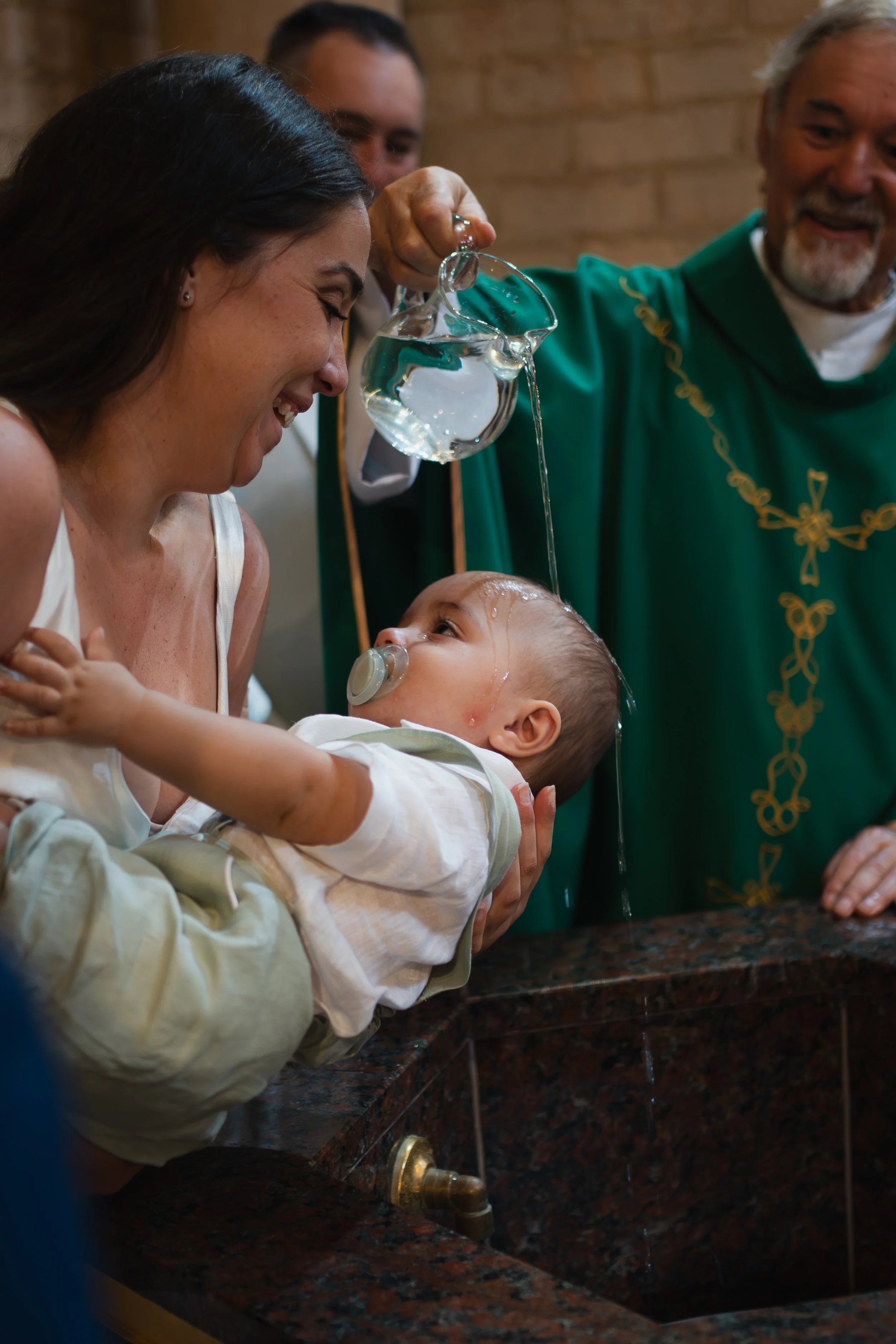 A baby being baptized, with water poured over their head by a priest, while a woman holds the baby and smiles.