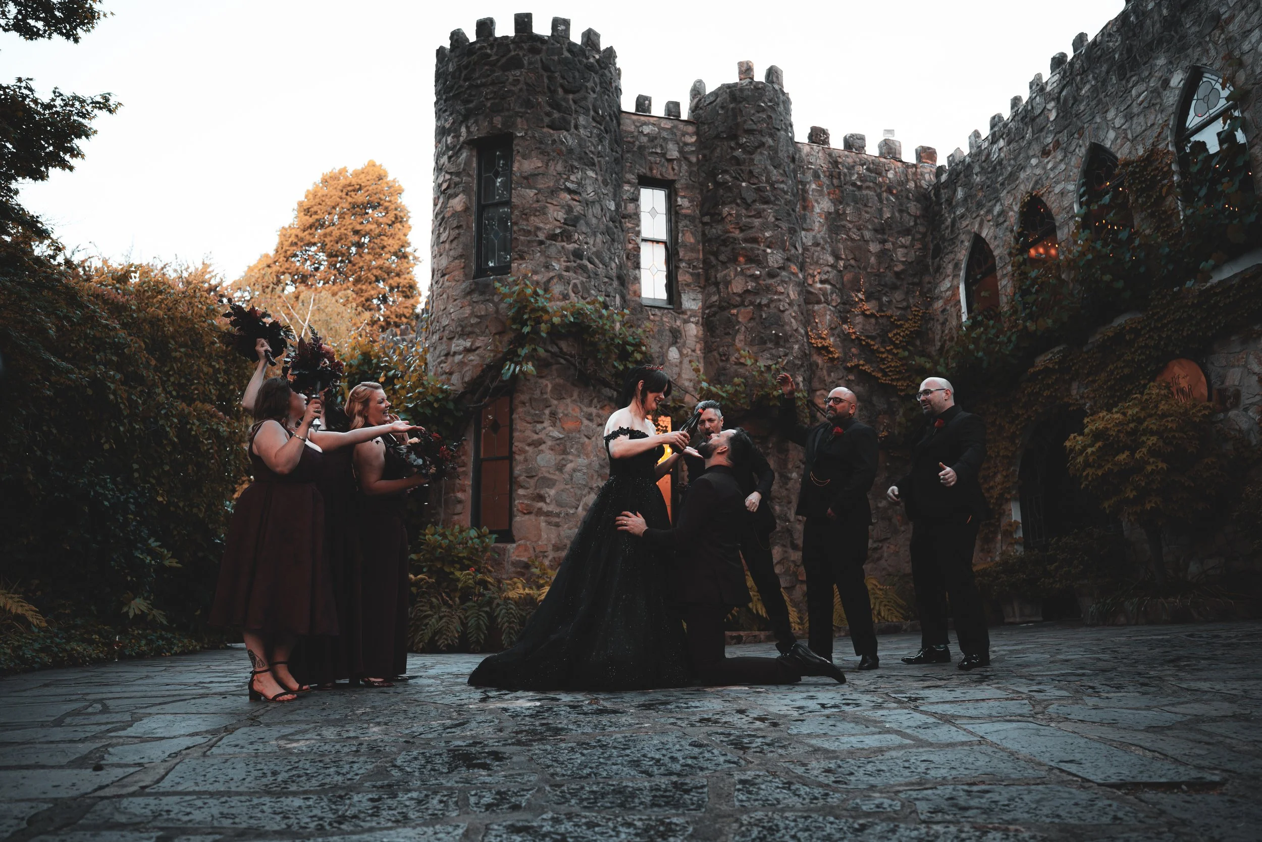 A wedding ceremony takes place outdoors in front of a stone castle-like building, with one woman kneeling and another woman pouring a drink for her, while officiating and four men and three women in formal attire look on.