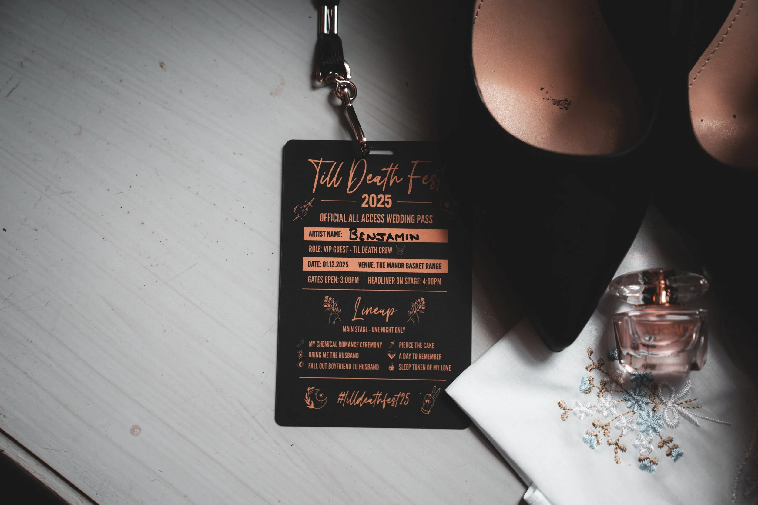 Wedding pass labeled 'Till Death Fest 2025' with artist name Benjamin, for VIP guest role, dated 01.12.2025, venue The Manor Basket Range, and a lineup for main stage. Alongside a perfume bottle, a black cup, and a embroidered white cloth.