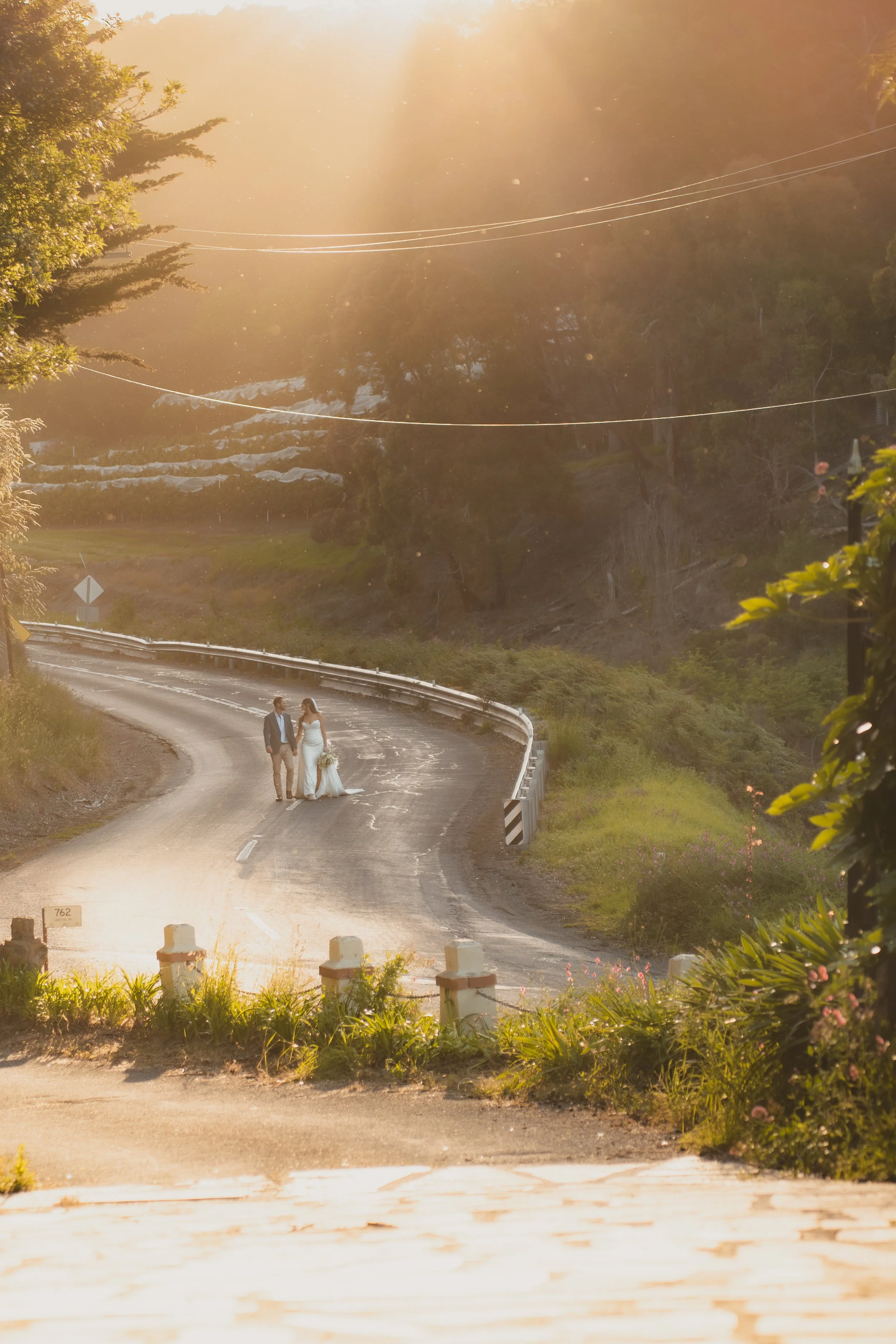 A bride and groom holding hands and walking on a winding road during sunset, surrounded by lush greenery.