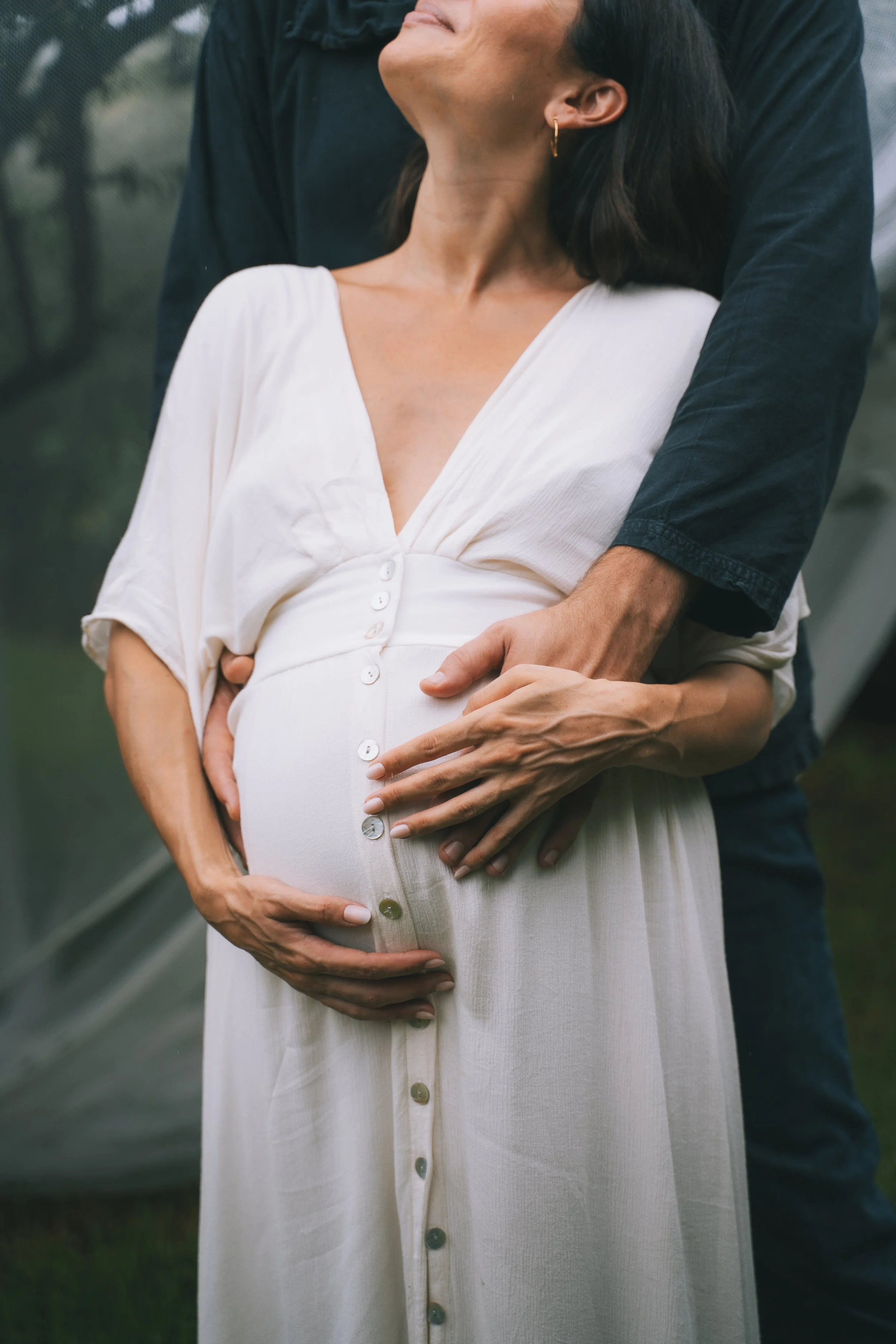 A pregnant woman in a white dress is being embraced by a man, with his arms around her belly, in an outdoor setting.