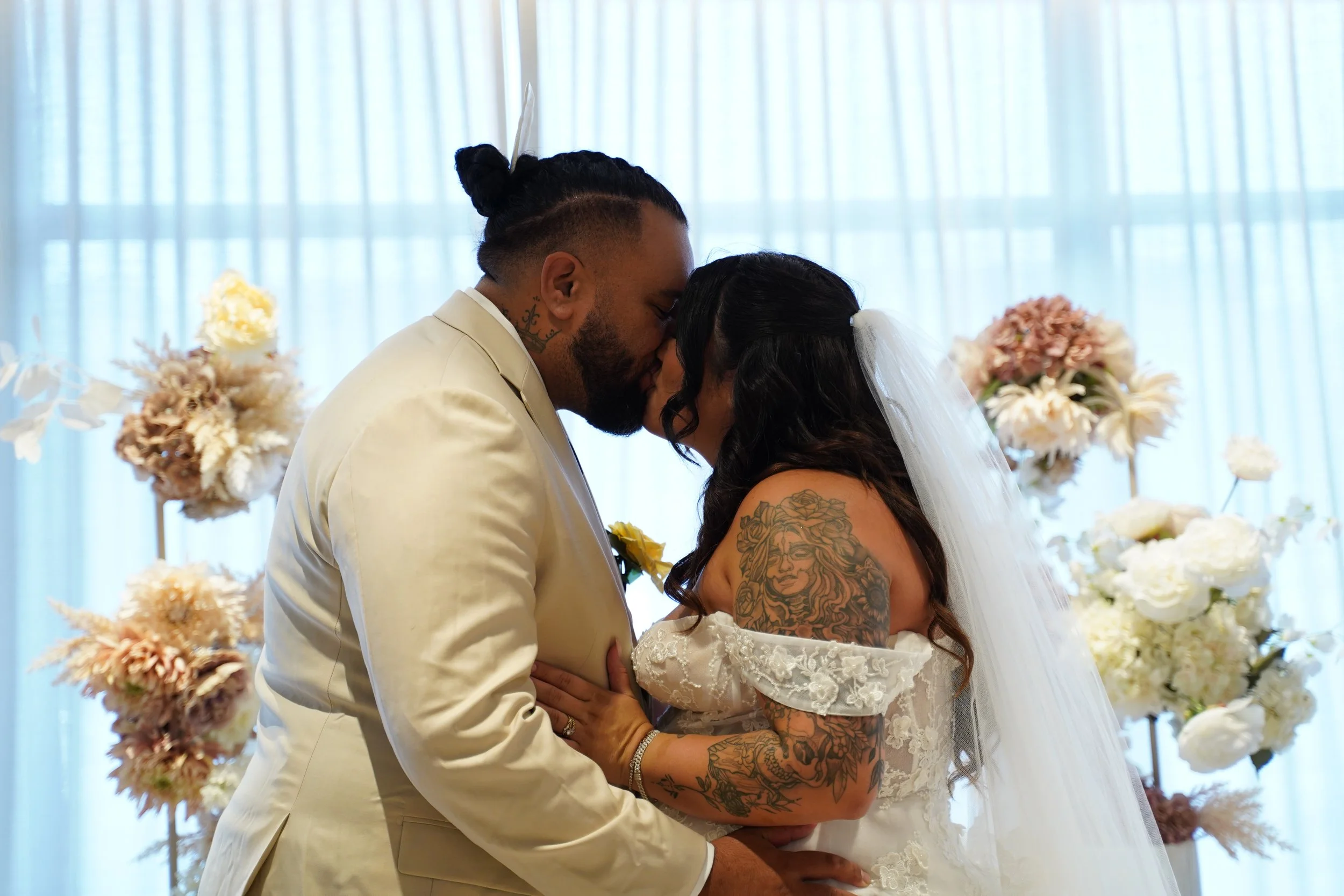 Couple in wedding attire sharing a kiss with floral arrangements and sheer curtains in the background.