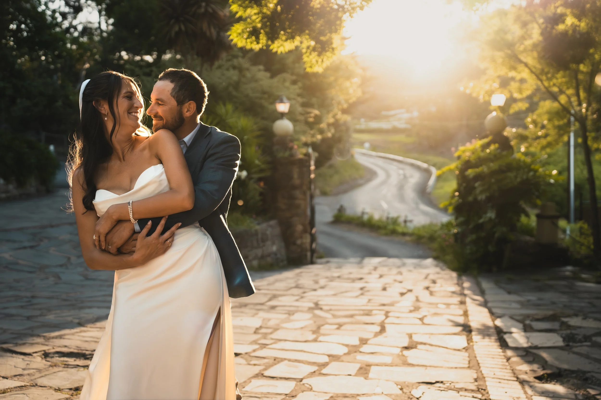A happy couple dressed in wedding attire sharing a joyful moment outdoors during sunset on a cobblestone path surrounded by lush greenery.