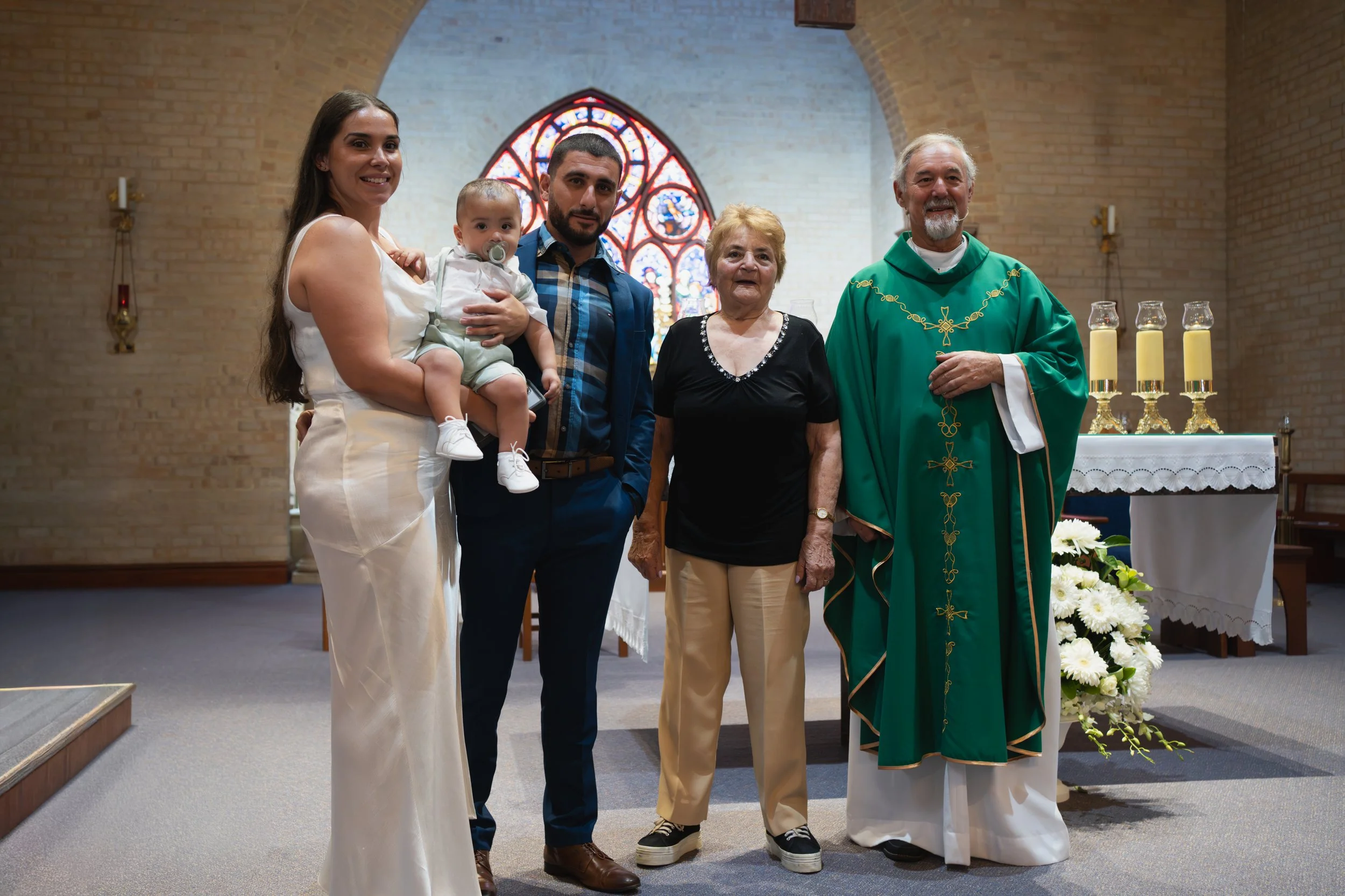 A family standing inside a church during a baptism ceremony. The woman on the left is holding a young child, and there's a man, an elderly woman, and a priest dressed in green robes. Stained glass window behind them. An altar with candles and flowers