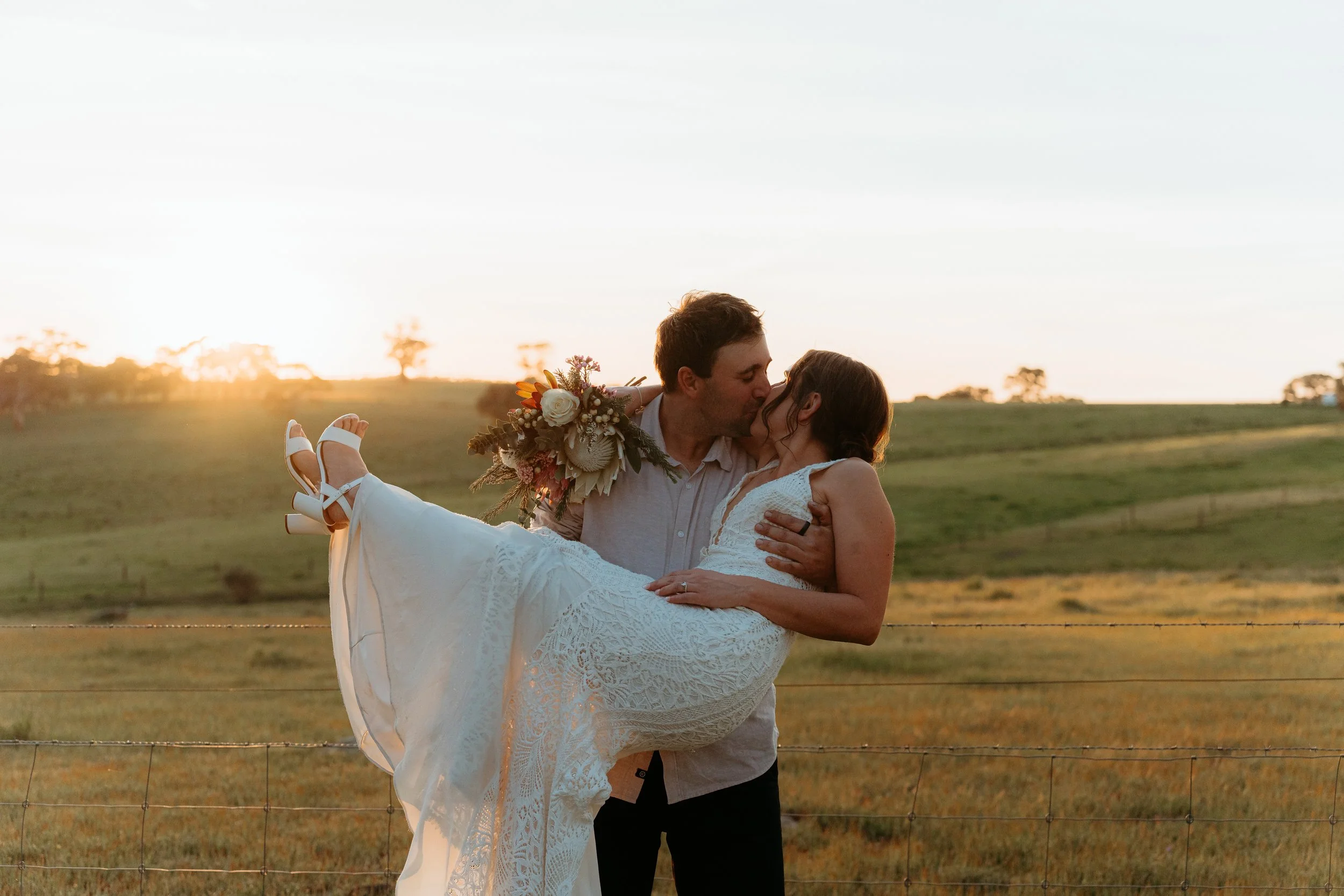 A couple embraces in a field during sunset, with the man holding the woman in a white dress and bouquet, sharing a kiss.