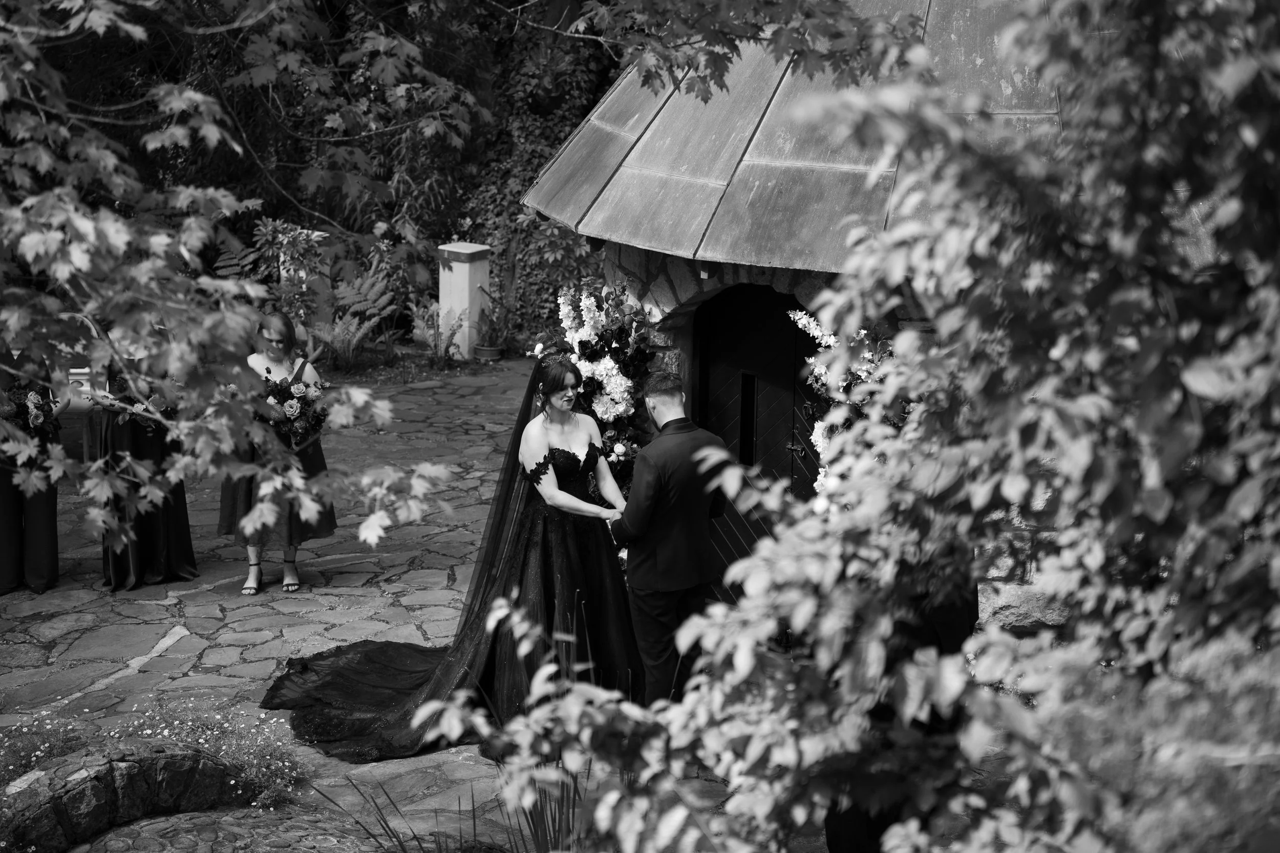 A black and white photo of a wedding ceremony outdoors, with a bride and groom holding hands in front of a small stone structure with a metal roof, surrounded by trees, flowers, and bridesmaids holding bouquets.